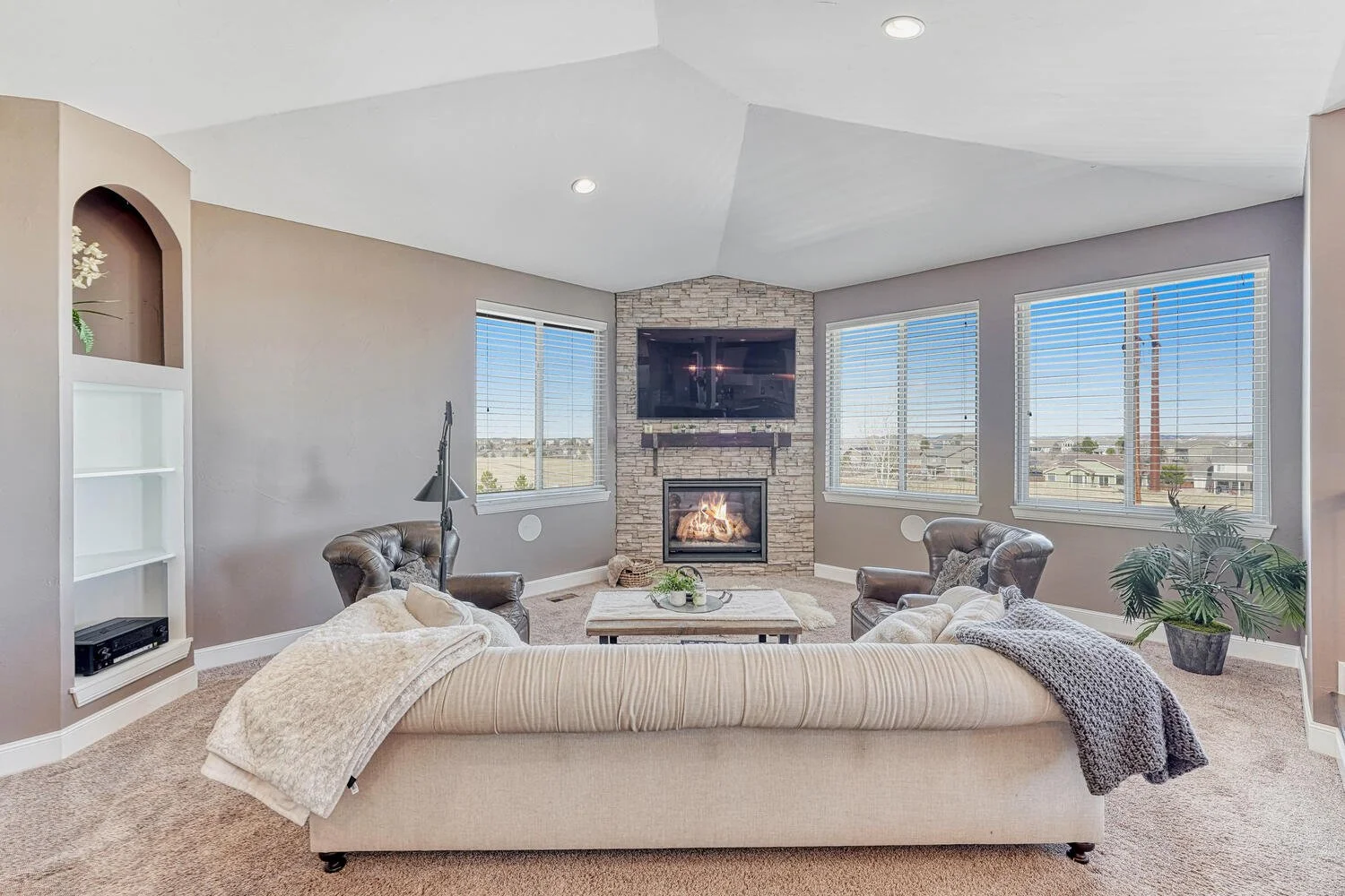 Inviting family room featuring a floor-to-ceiling stone fireplace, wall-mounted television, vaulted ceiling, large windows, and comfortable seating