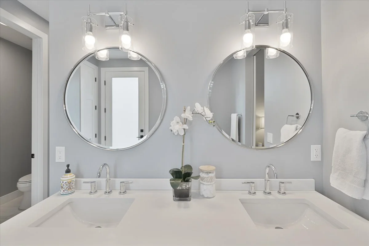 Custom white bathroom vanity featuring dual undermount sinks, polished chrome faucets, round framed mirrors, and contemporary vanity lighting