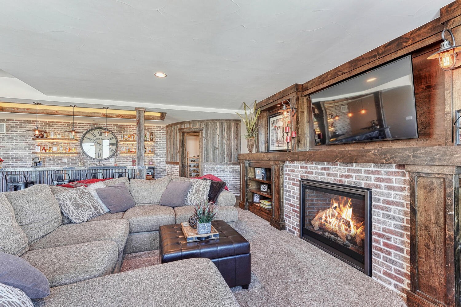 Rustic finished basement living area featuring a brick fireplace, reclaimed wood accents, large sectional sofa, built-in entertainment center, and custom bar with illuminated shelving.
