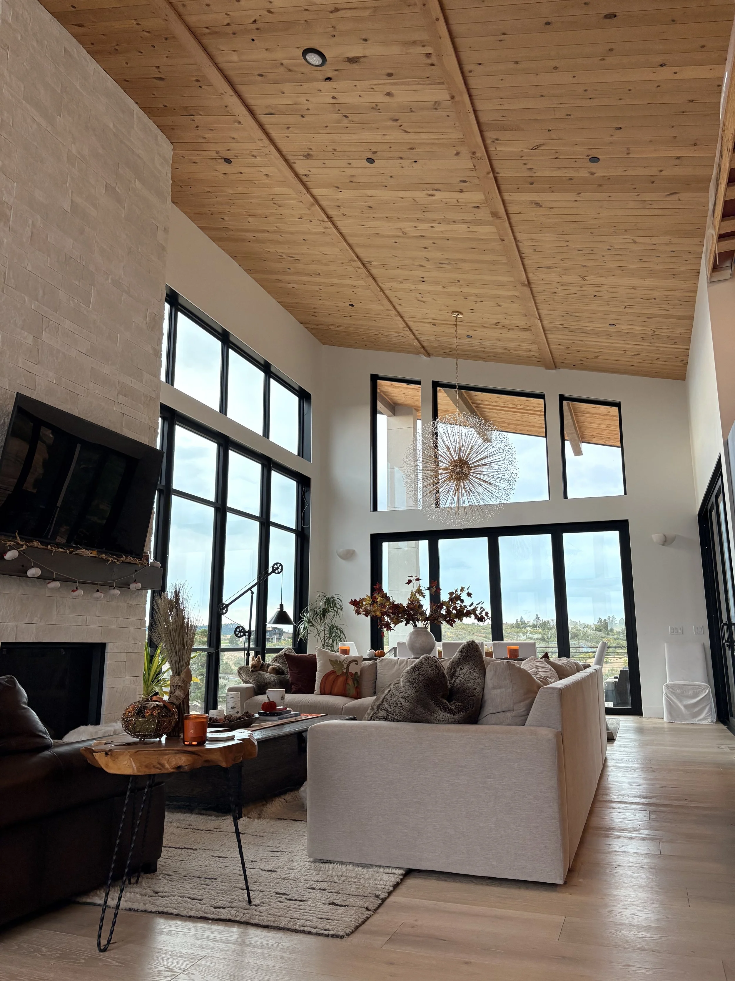 Spacious great room featuring a vaulted wood plank ceiling, floor-to-ceiling black-framed windows, stone fireplace, modern chandelier, and neutral furnishings.