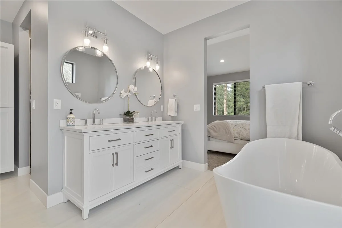 Elegant primary bathroom featuring a white double-sink vanity, round mirrors, chrome lighting fixtures, freestanding soaking tub, and soft gray walls