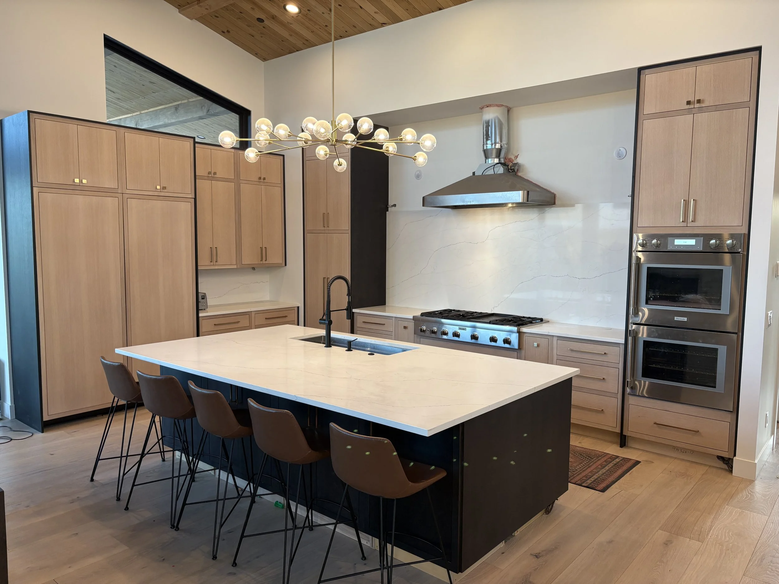 Contemporary kitchen featuring light wood cabinets, a white stone island with black base, stainless steel appliances, and modern chandelier lighting