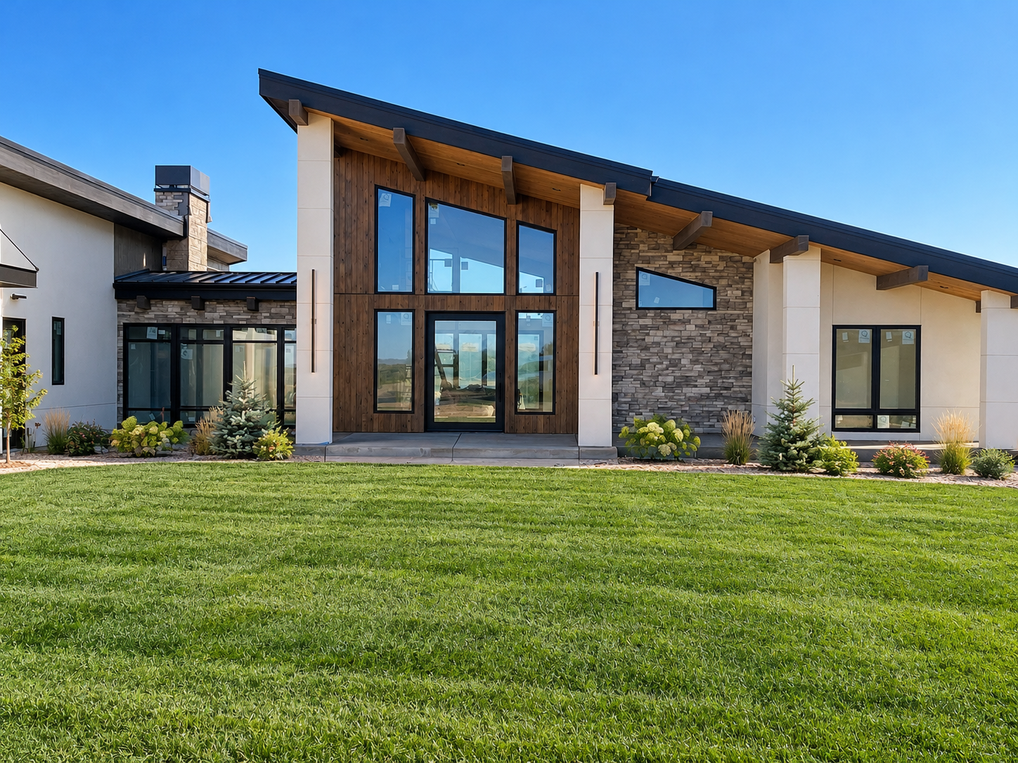 Modern custom home exterior featuring a dramatic sloped roofline, natural stone and wood accents, black-framed windows, a spacious entryway, and lush green lawn with landscaped garden bedsmodern custom home by Kloster Construction