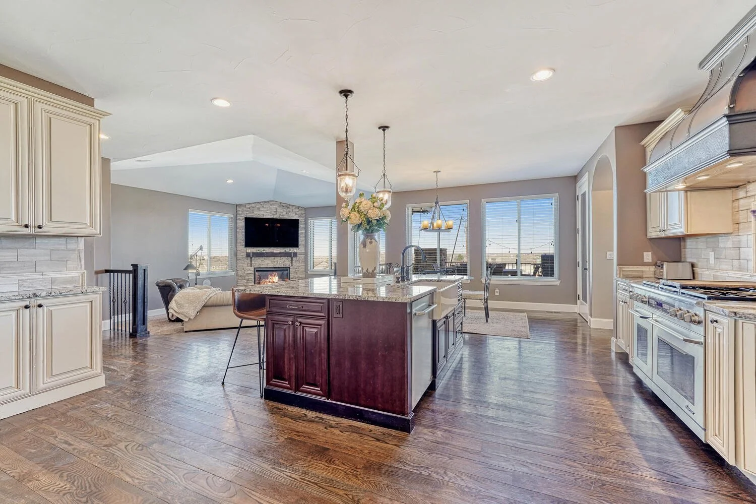 open-concept kitchen featuring cream-colored cabinetry, a dark wood island with granite countertops, stainless steel appliances, hardwood floors, and an adjoining living room with a stone fireplace.