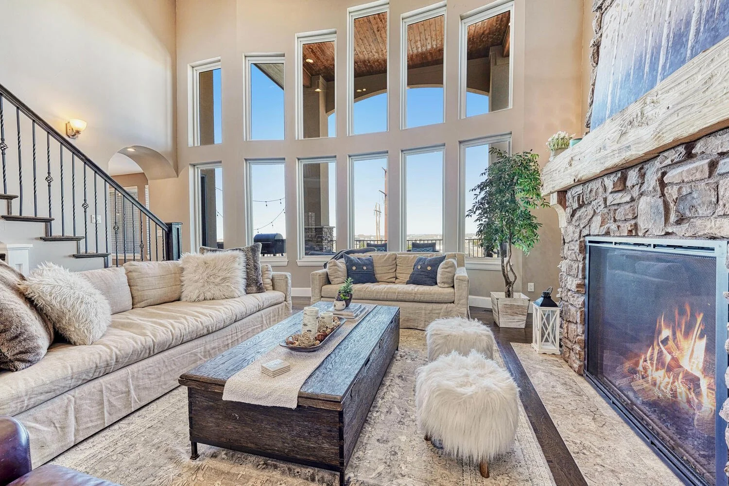 Two-story living room featuring a large natural stone fireplace, tall windows, hardwood flooring, neutral sofas, and a rustic wood coffee table.