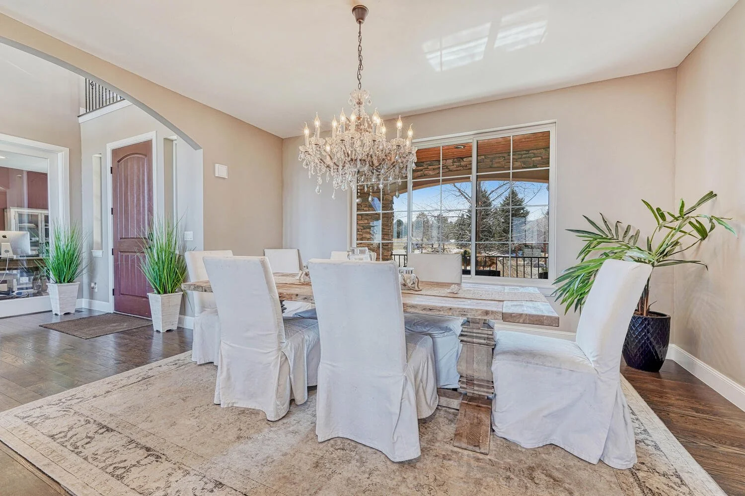Elegant dining room featuring a rustic wood table, upholstered white chairs, crystal chandelier, hardwood flooring, and a large window overlooking the patio.
