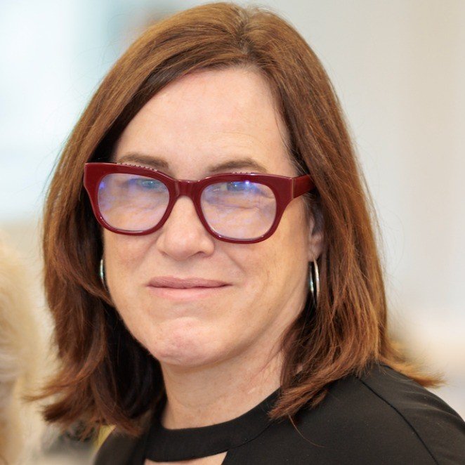 A woman with reddish-brown hair wearing red glasses and hoop earrings.
