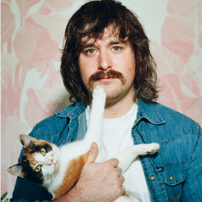 A man with long brown hair and a mustache holding a calico cat with one paw raised near his face.