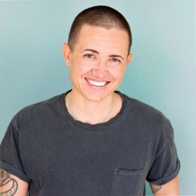 Smiling person with short hair wearing a dark t-shirt against a light blue background.