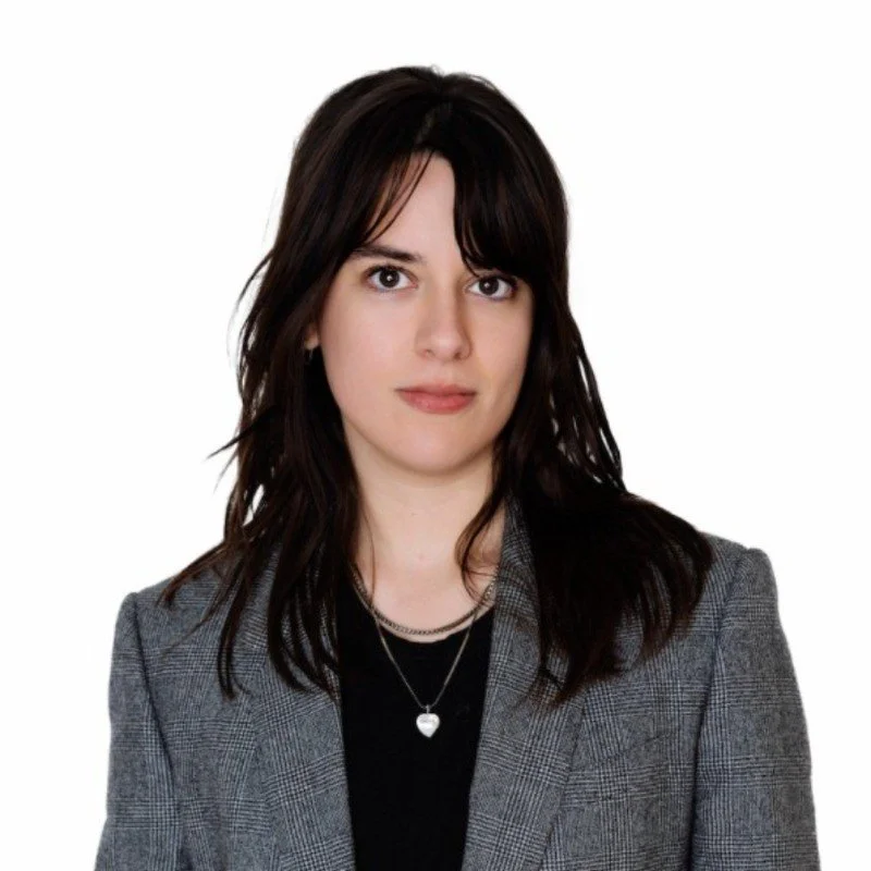 Portrait of a woman with shoulder-length dark brown hair, wearing a grey blazer over a black top, and a silver necklace with a heart pendant, against a white background.