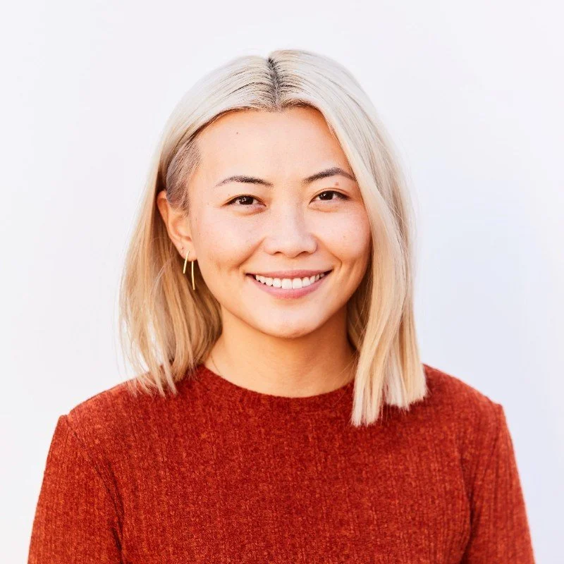 A smiling woman with shoulder-length blonde hair, wearing a red sweater and small hoop earrings, standing against a plain white background.