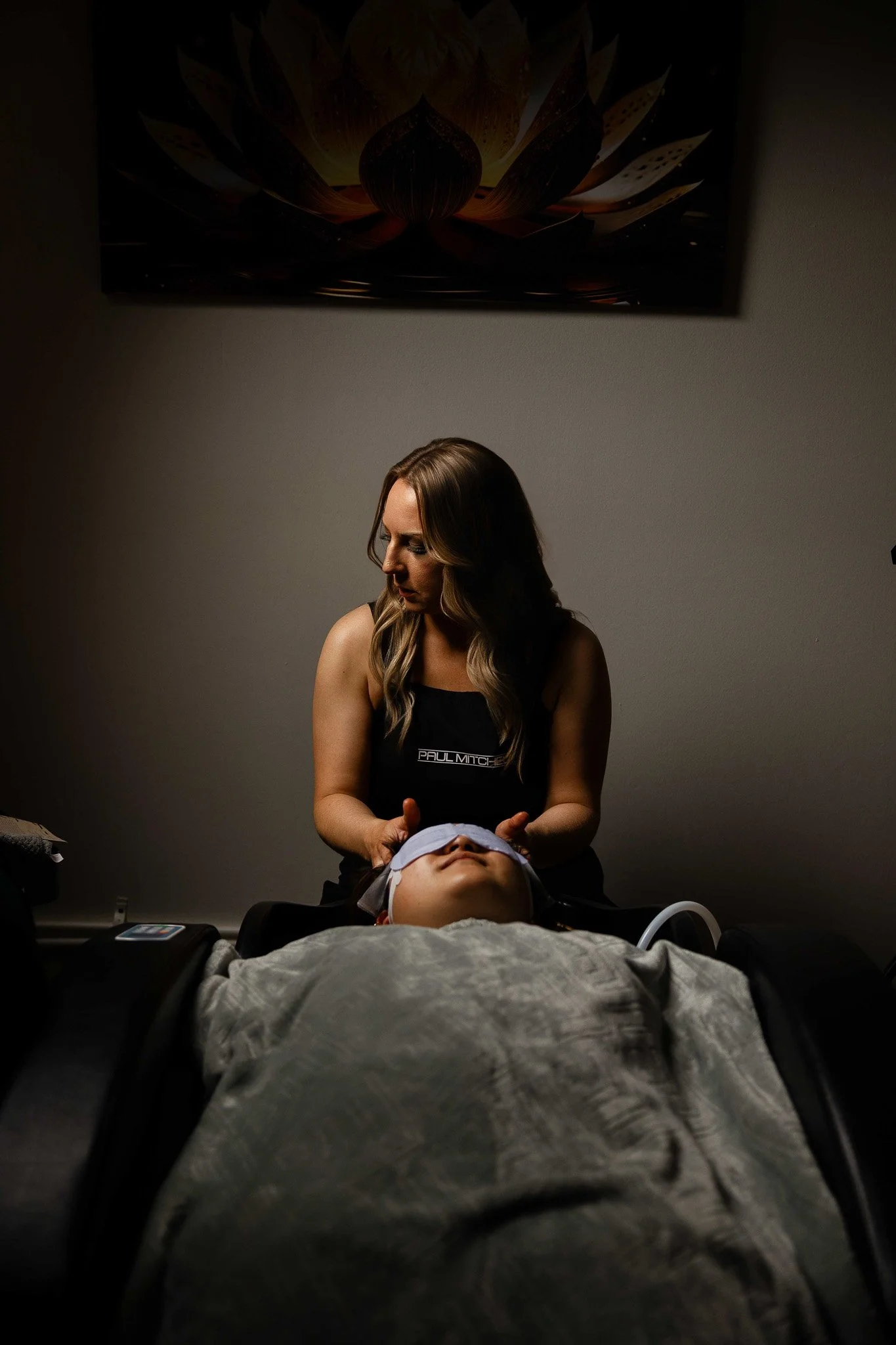 A woman is performing a facial massage or skincare treatment on a person lying down with their eyes closed, in a dimly lit room, with a flower painting on the wall behind them.