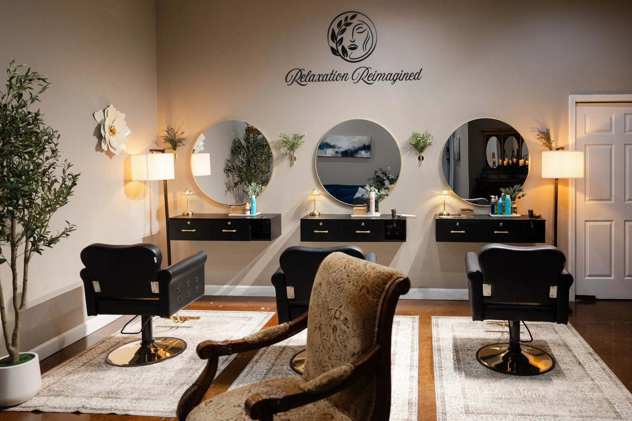 A relaxation spa room with three black vanity tables each with a mirror, small lamps, and skincare products on top. There are four black chairs in front of the tables and an upholstered armchair in the foreground. A large plant is on the left and inside the mirrors, reflections of more plants and a painting can be seen. The wall has a logo of a face with a Laurel wreath and the words "Relaxation Reimagined."