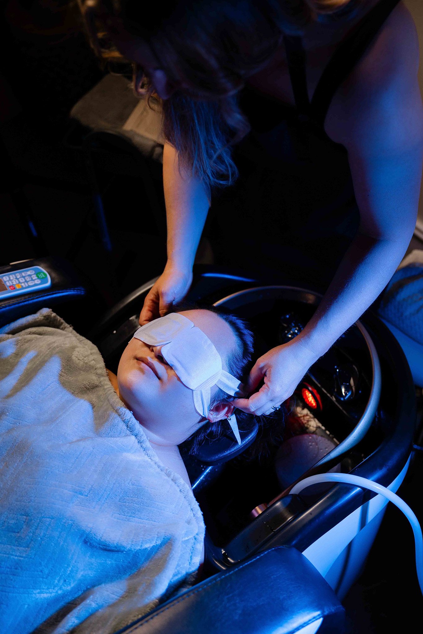 A woman with a bandage on her forehead lies inside an ambulance or emergency vehicle, while another woman leans over her, tending to her with care. The scene is dimly lit with blue and red emergency lights.
