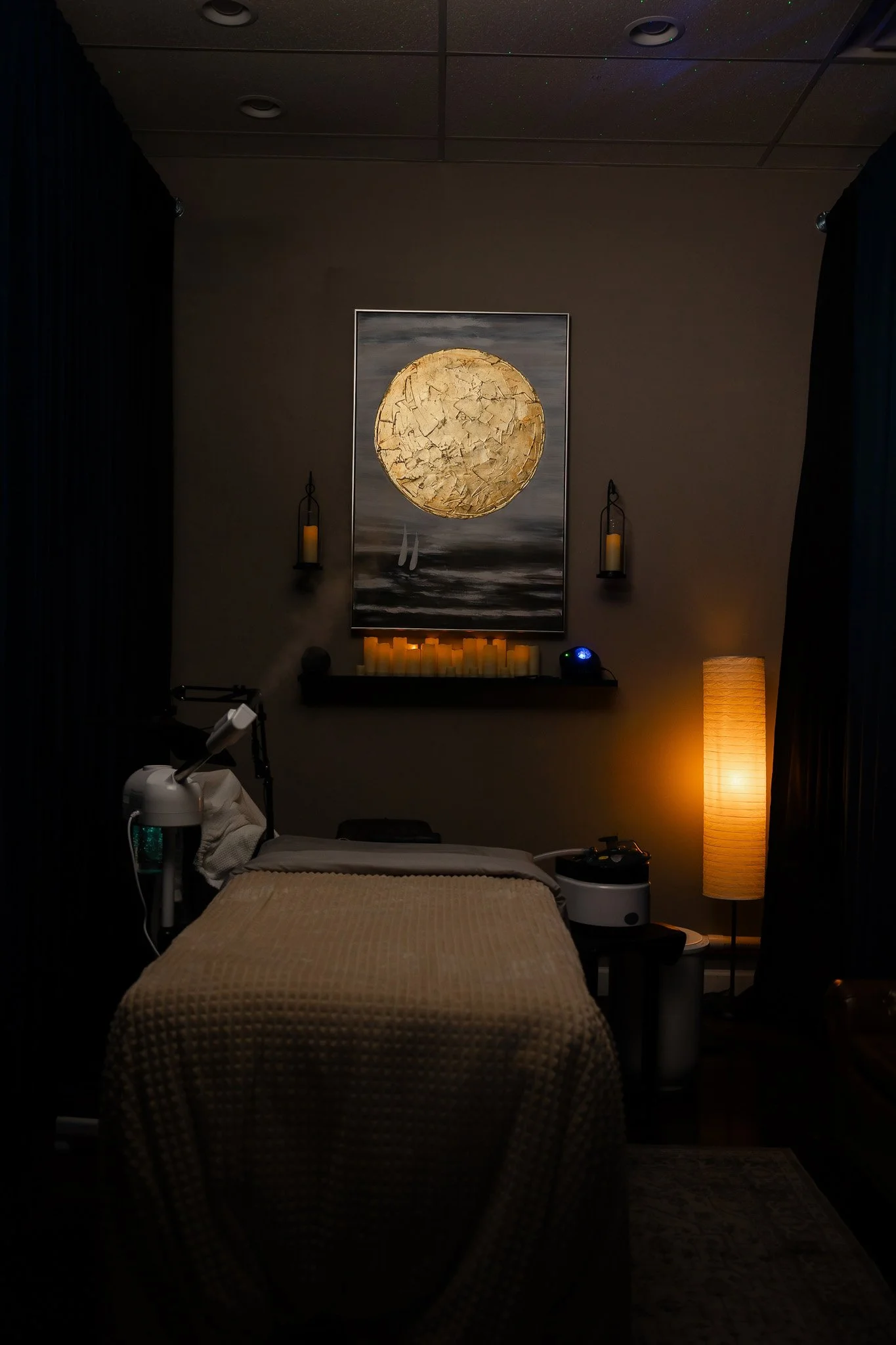 Dimly lit room with a massage table covered in a textured beige blanket, surrounded by soft lighting including candles, a floor lamp, and wall sconces, with a large abstract artwork featuring a gold circle on the wall.