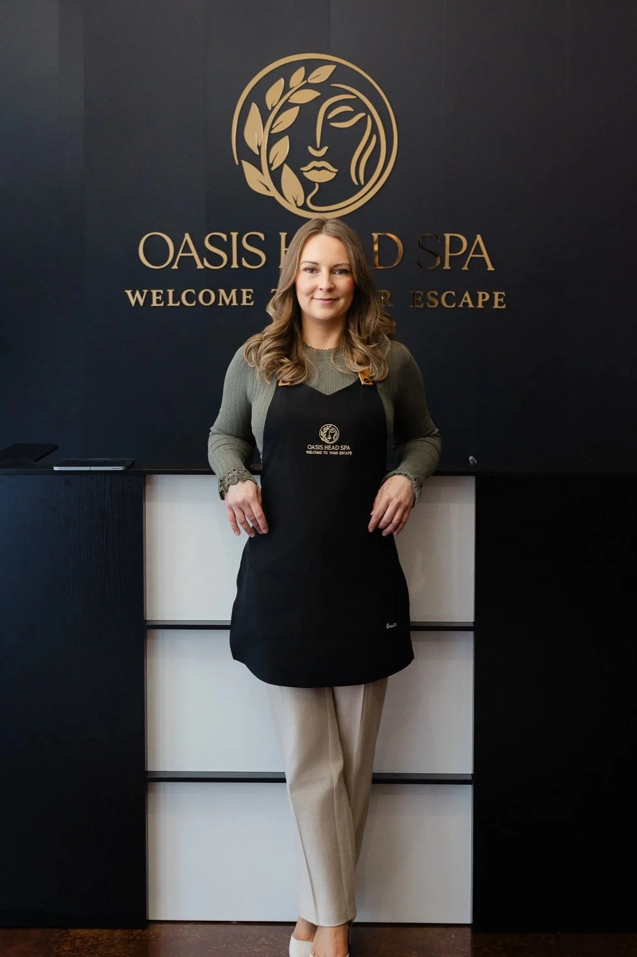 A woman with long brown hair wearing a green sweater and beige pants stands in front of a black wall with gold text and logo for Oasis Head Spa. She is smiling, wearing an apron with the spa's logo, and standing with her hands on her hips.