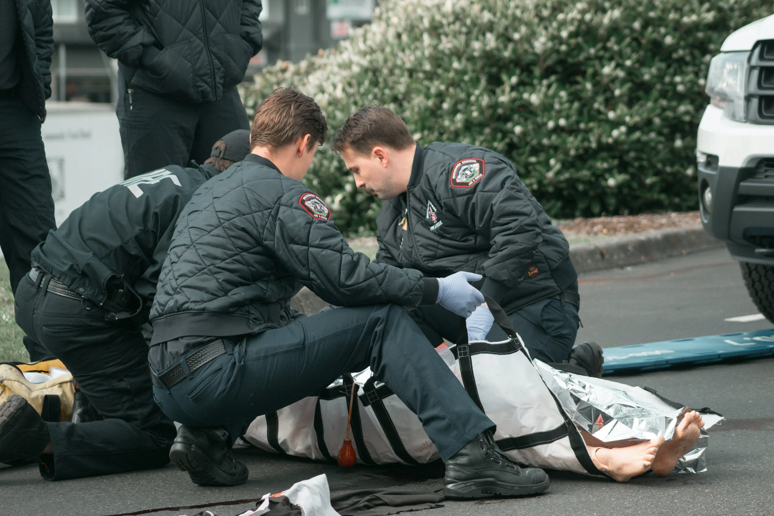 Emergency medical personnel attending to a person lying on the ground, who appears to be injured or unconscious, on a city street with bushes and vehicles in the background.