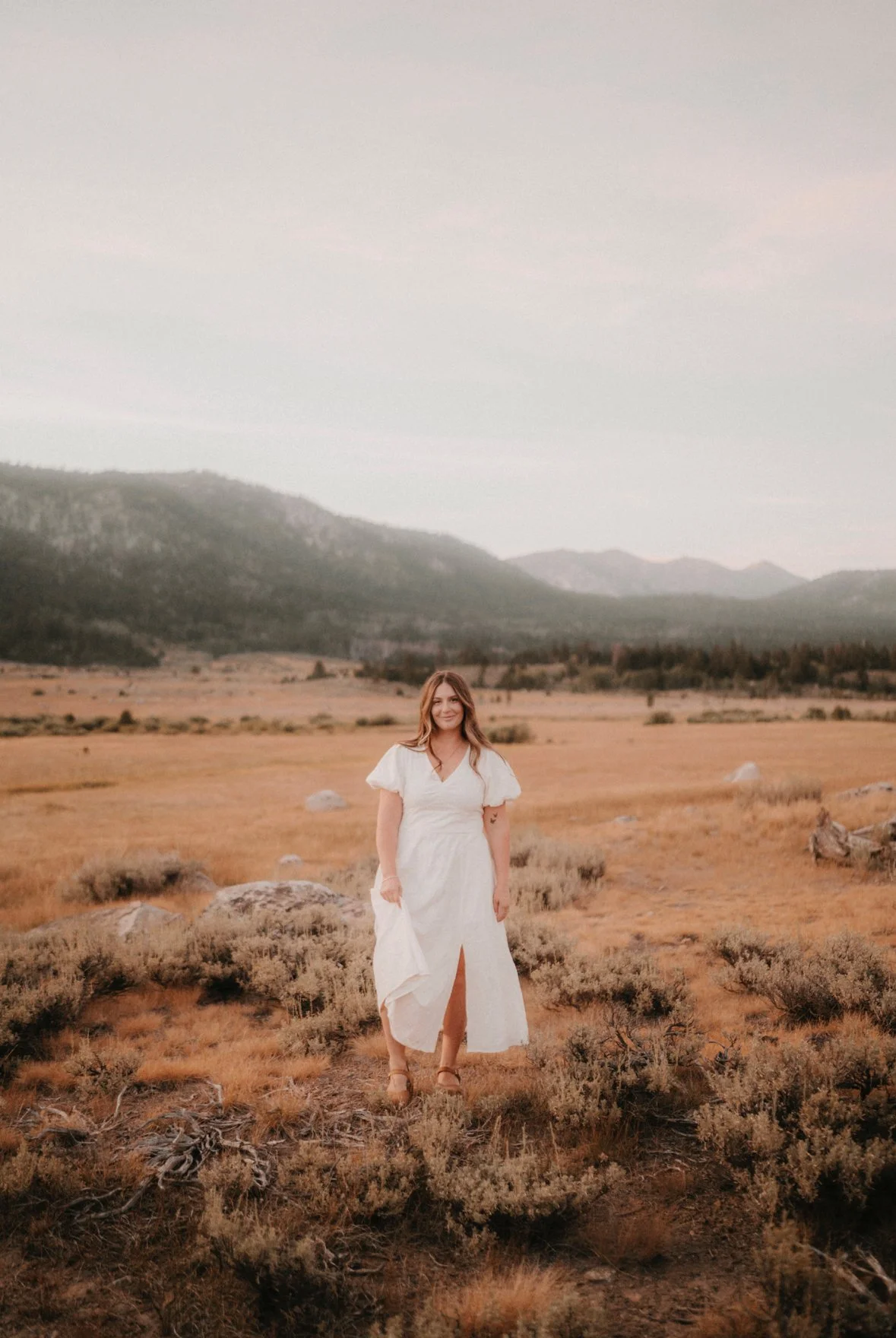 A woman in a white dress standing in a desolate field with mountains in the background during sunset.