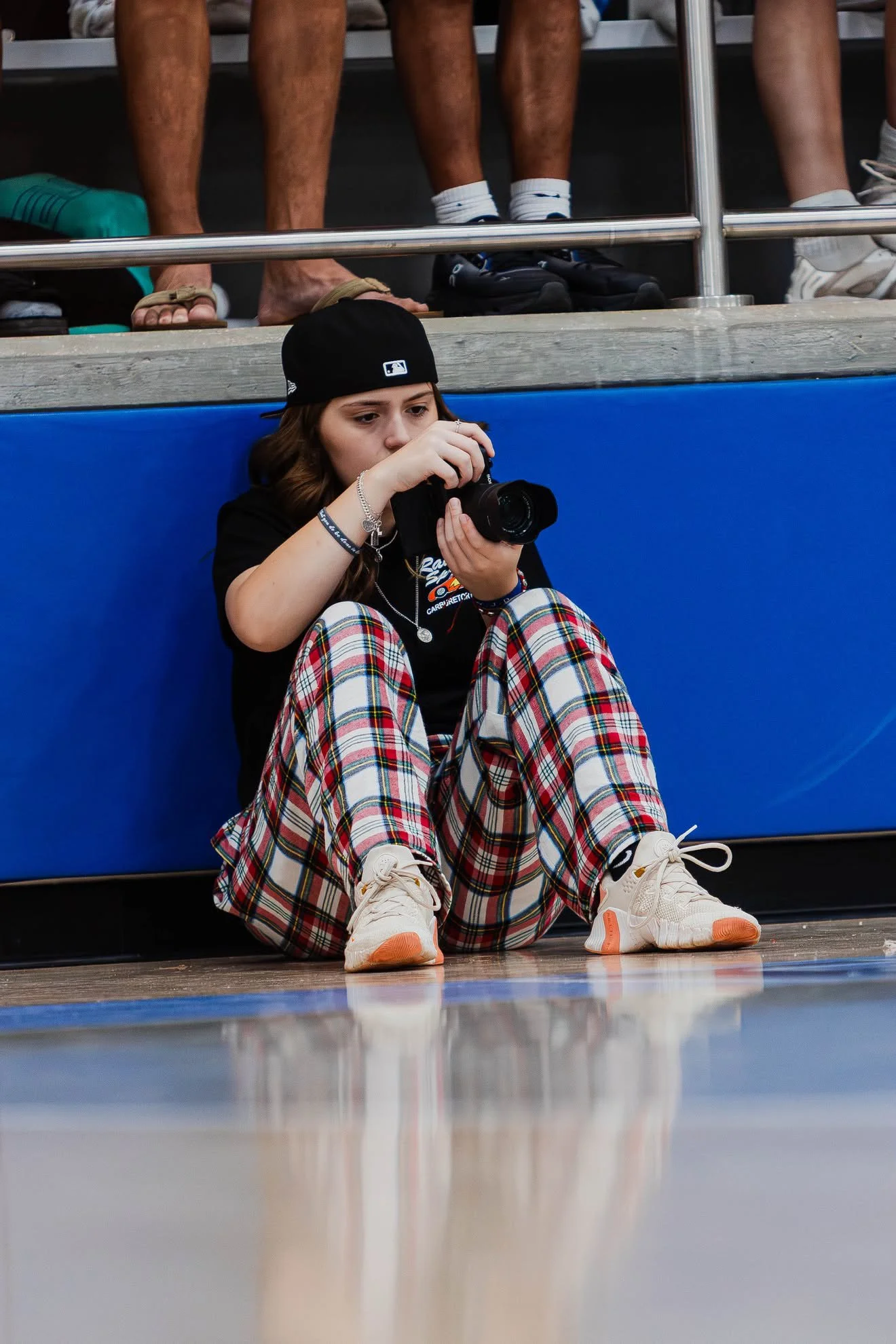 A young woman with long hair, wearing a black baseball cap, black shirt, plaid pants, and beige sneakers, is sitting on the floor, holding a camera, and looking at it. In the background, people are sitting on a bench, with their legs visible.