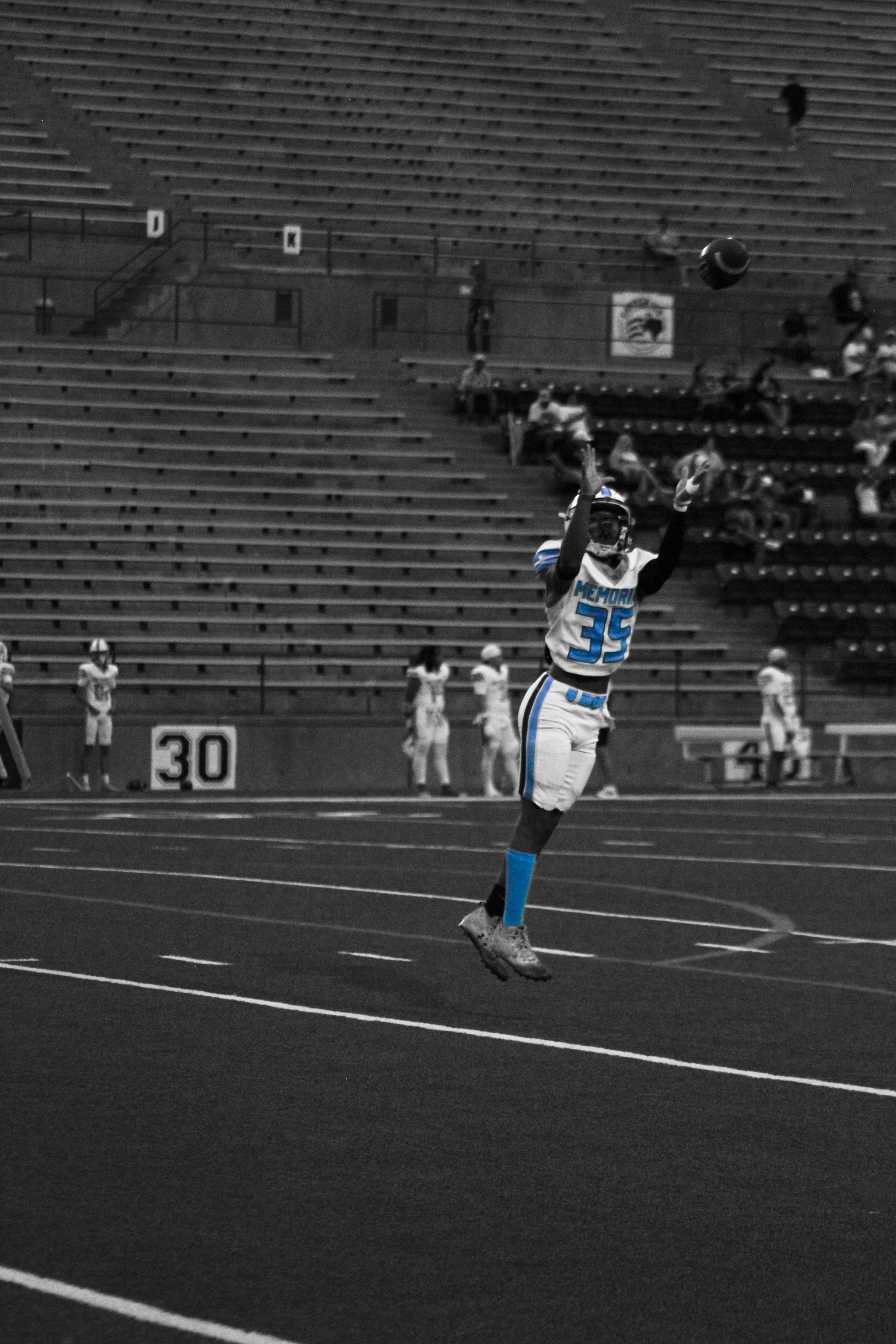 A football player in a white and blue uniform, numbered 35, is jumping in the air to catch a football during a game. The stadium seats are mostly empty, with a few spectators visible in the background.