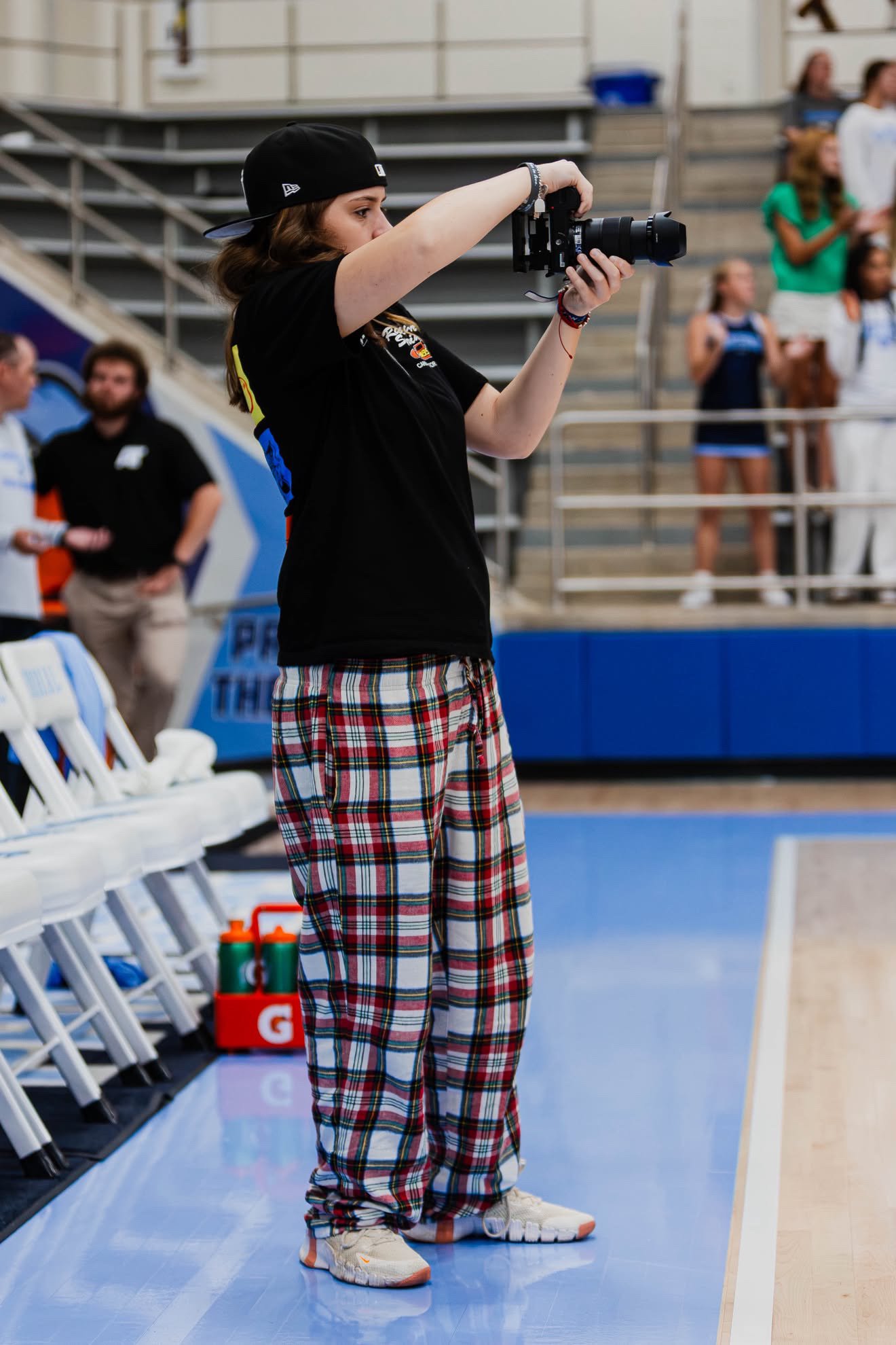 A young woman with a black hat and plaid pants taking photographs with a camera at a sports event. In the background, other spectators are watching, and the setting appears to be an indoor stadium or arena.