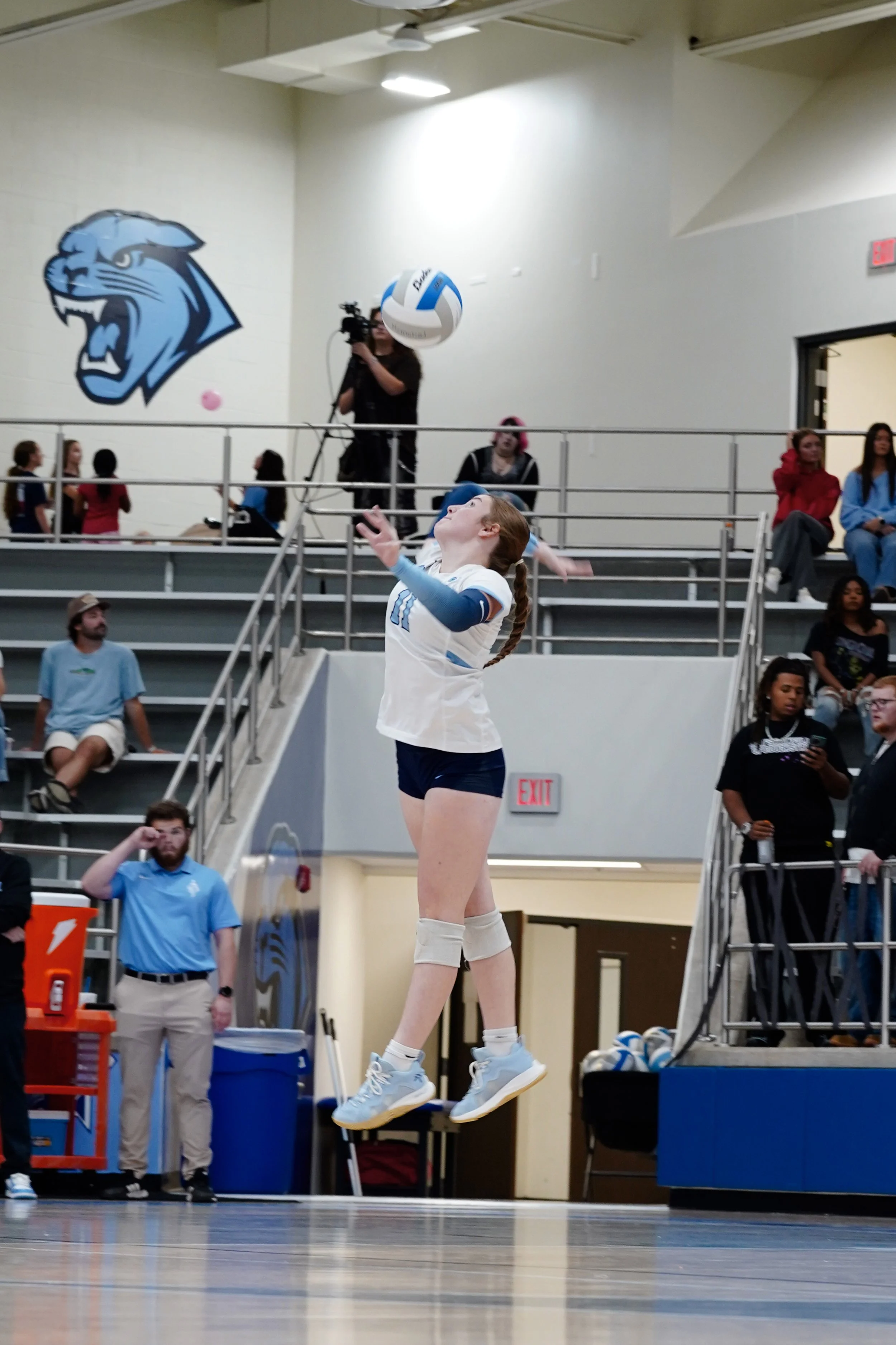 A female volleyball player in mid-air preparing to hit the ball during a game at a gymnasium. The player is wearing a white uniform with blue accents, knee pads, and sneakers. Spectators and officials are visible in the background, along with a large blue and white volleyball and a wall with a lion mascot logo.