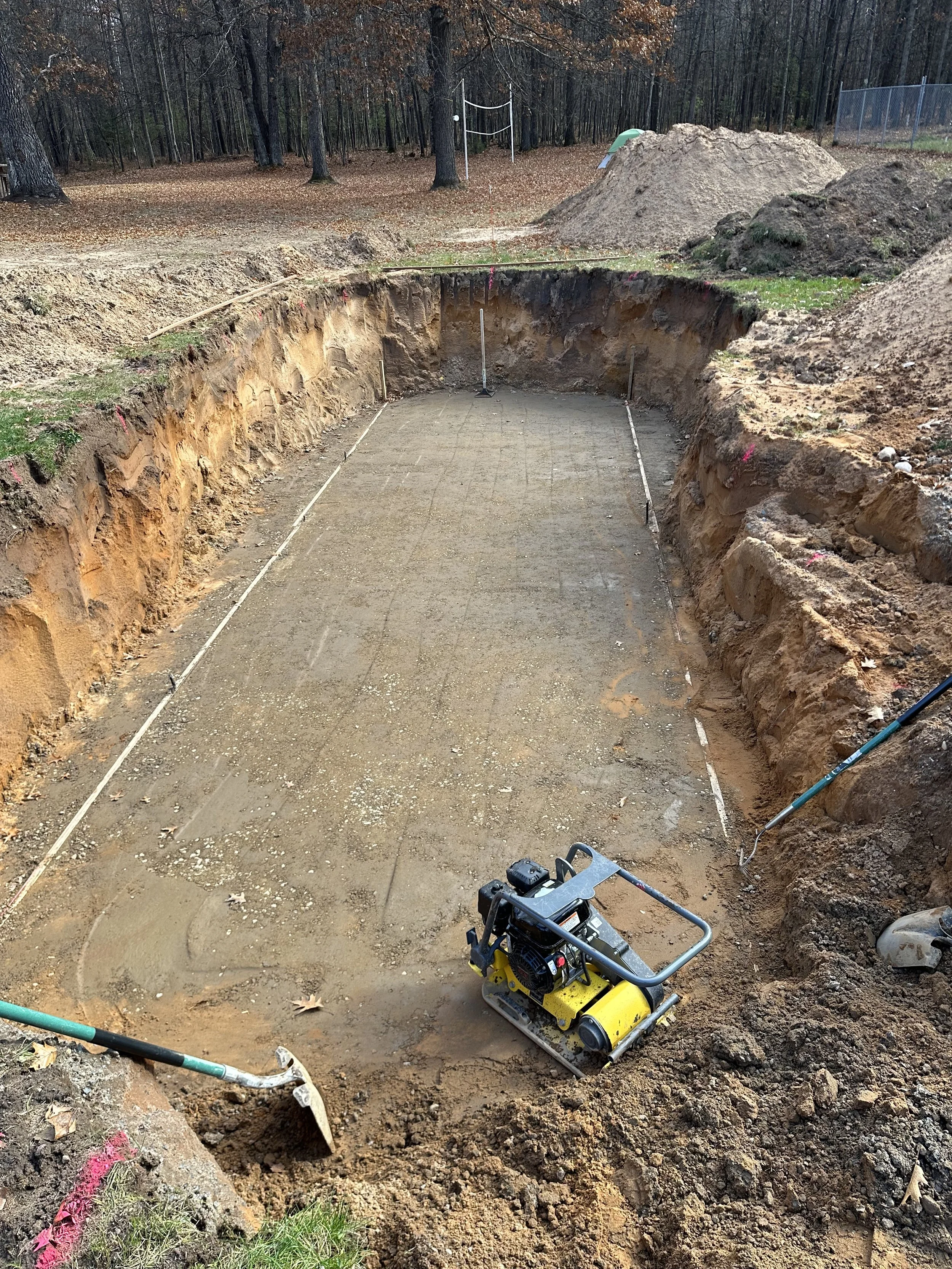 Excavated rectangular foundation site in a wooded backyard, with a yellow plate compactor, soil piles, utility markings, and a volleyball net.