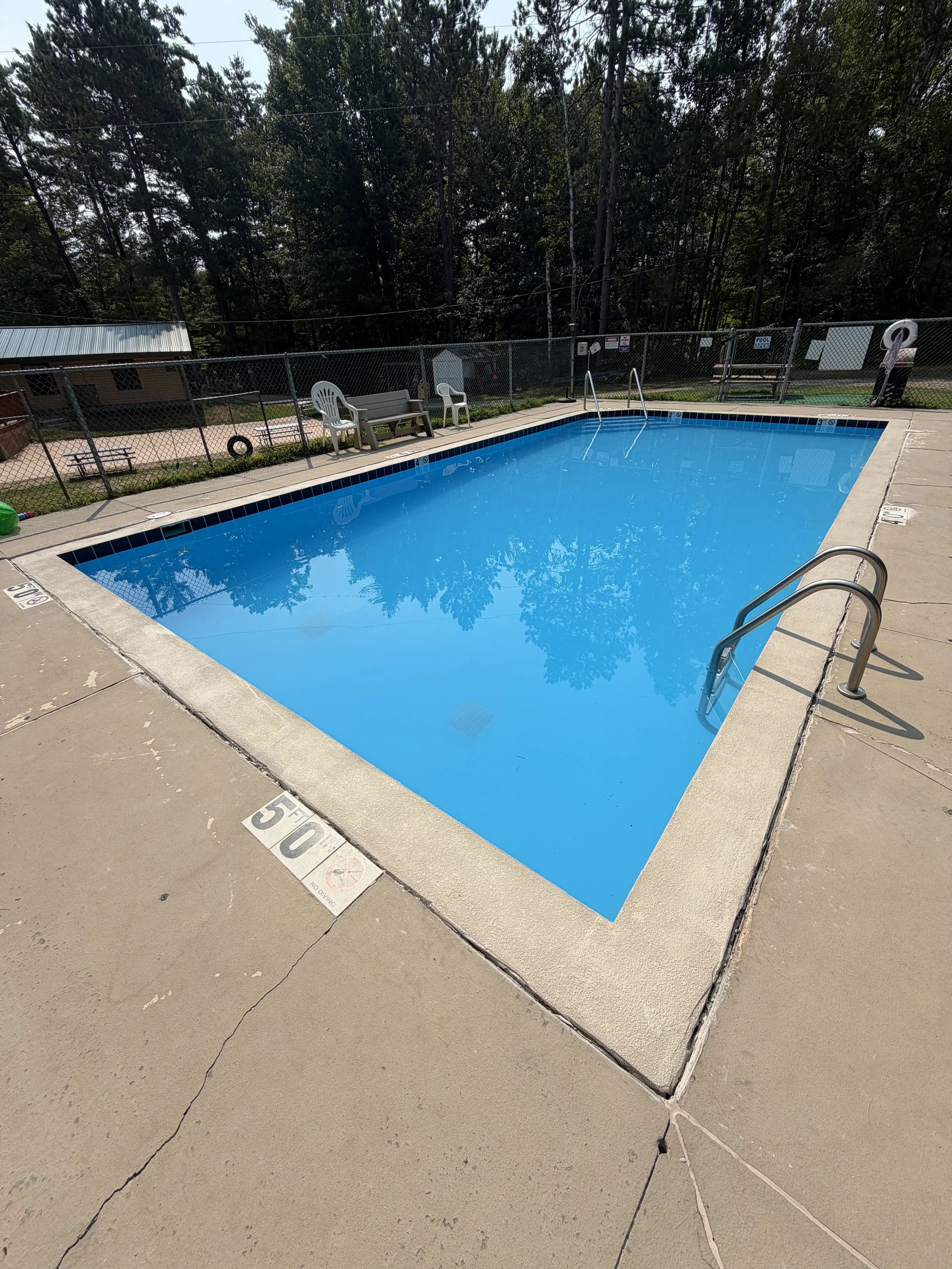 Outdoor swimming pool with diving-depth markers, metal ladders, lounge chairs, safety fencing, and wooded surroundings