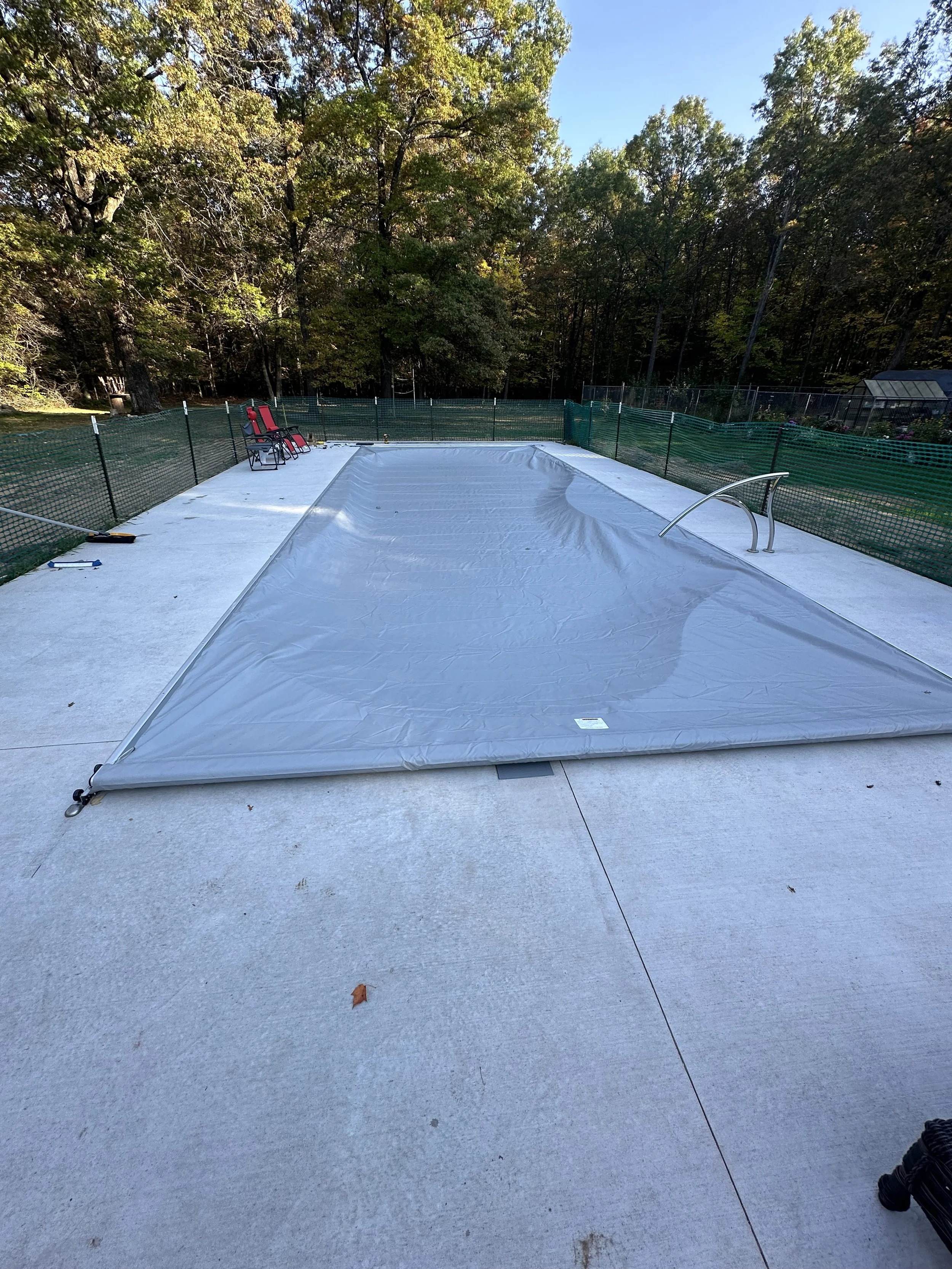 Covered outdoor swimming pool surrounded by green safety fencing, lounge chairs, and wooded autumn landscape