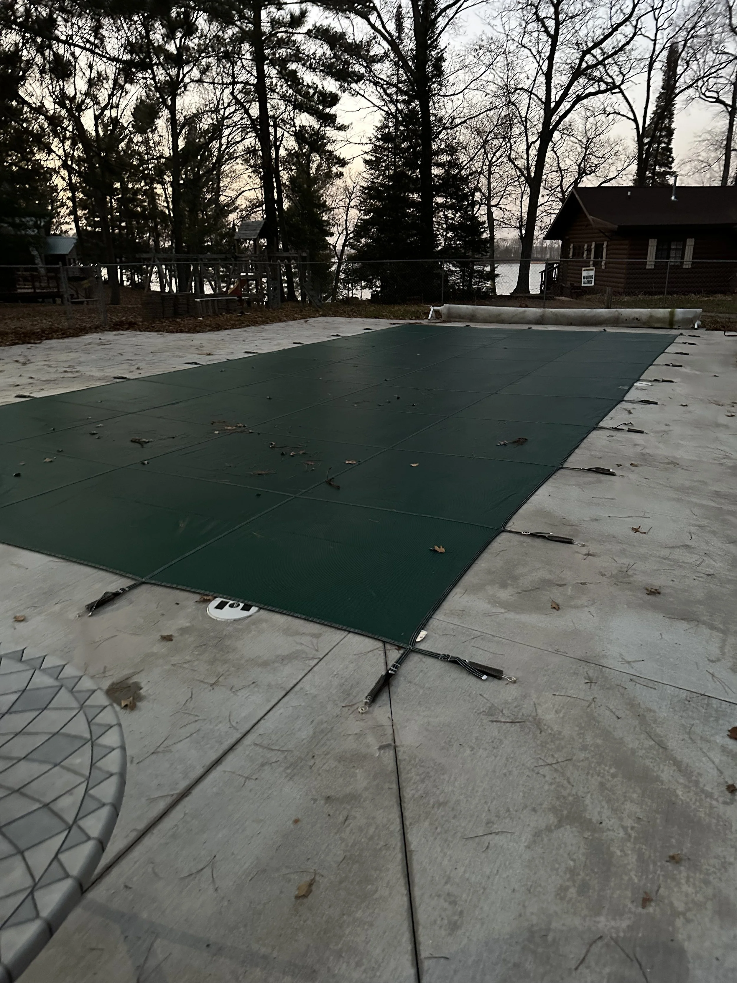 Outdoor pool covered with dark green panels beside a wooded log cabin and chain-link fence.
