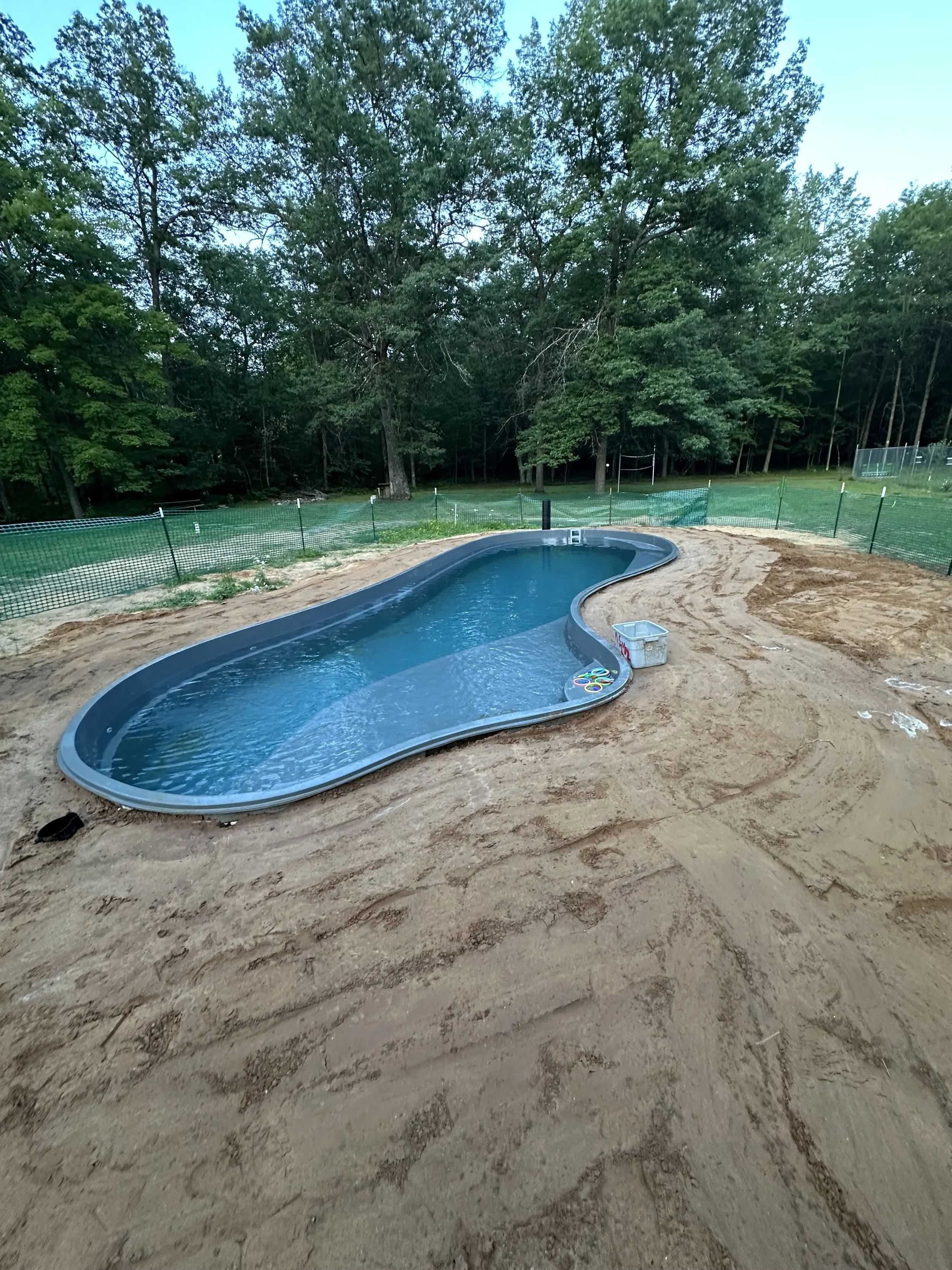 Newly installed in-ground swimming pool surrounded by sandy soil, temporary fencing, and trees.