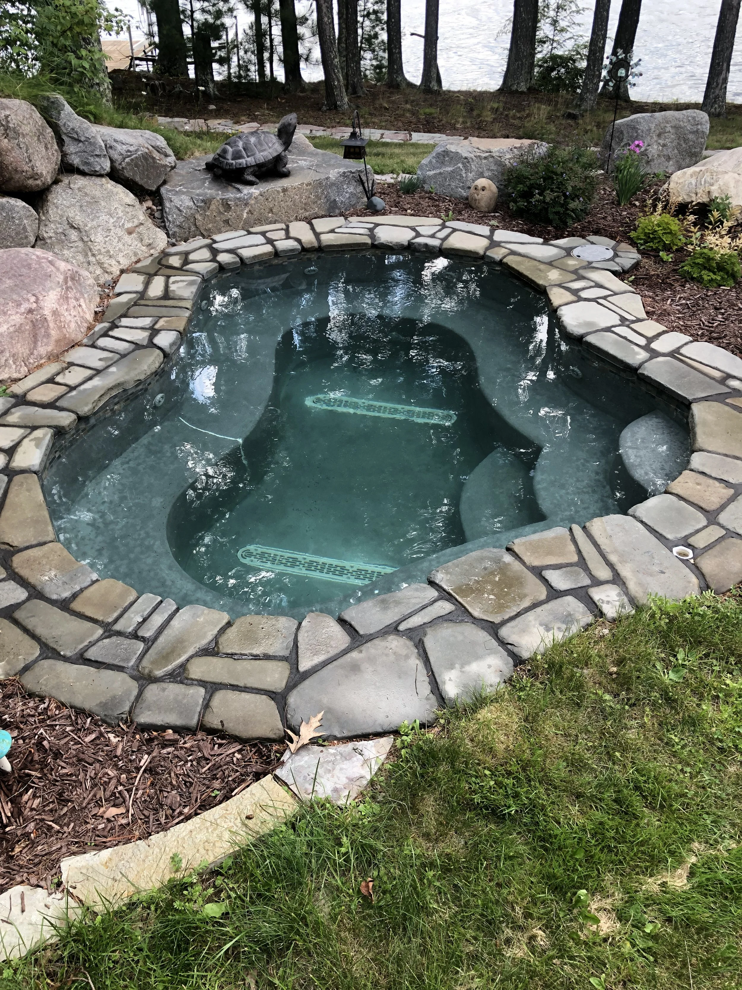 Stone-surrounded backyard hot tub beside a garden, with turtle and owl ornaments, plants, rocks, and trees overlooking a lake.