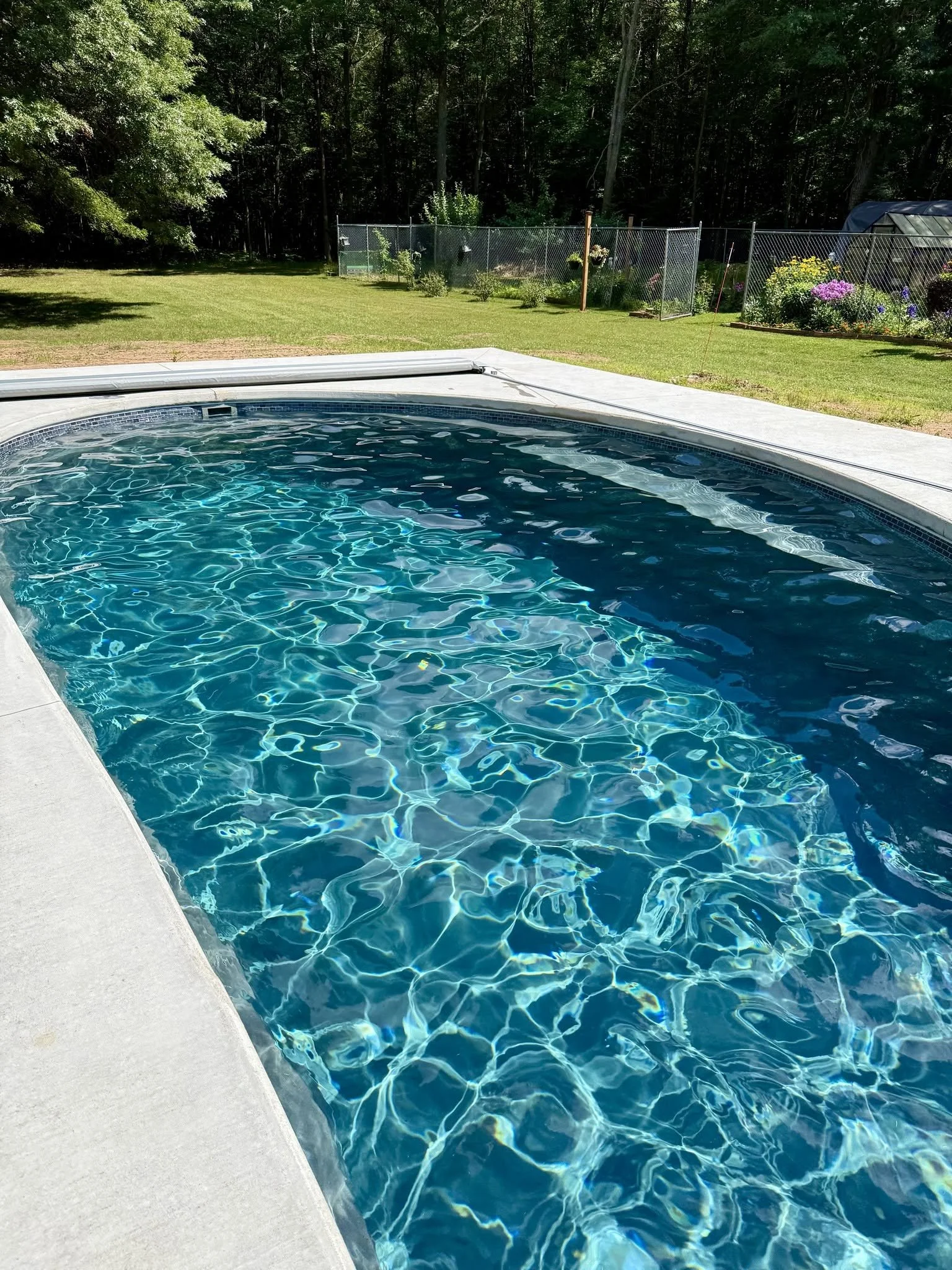 Backyard swimming pool with shimmering blue water, lawn, flower garden, and wooded backdrop