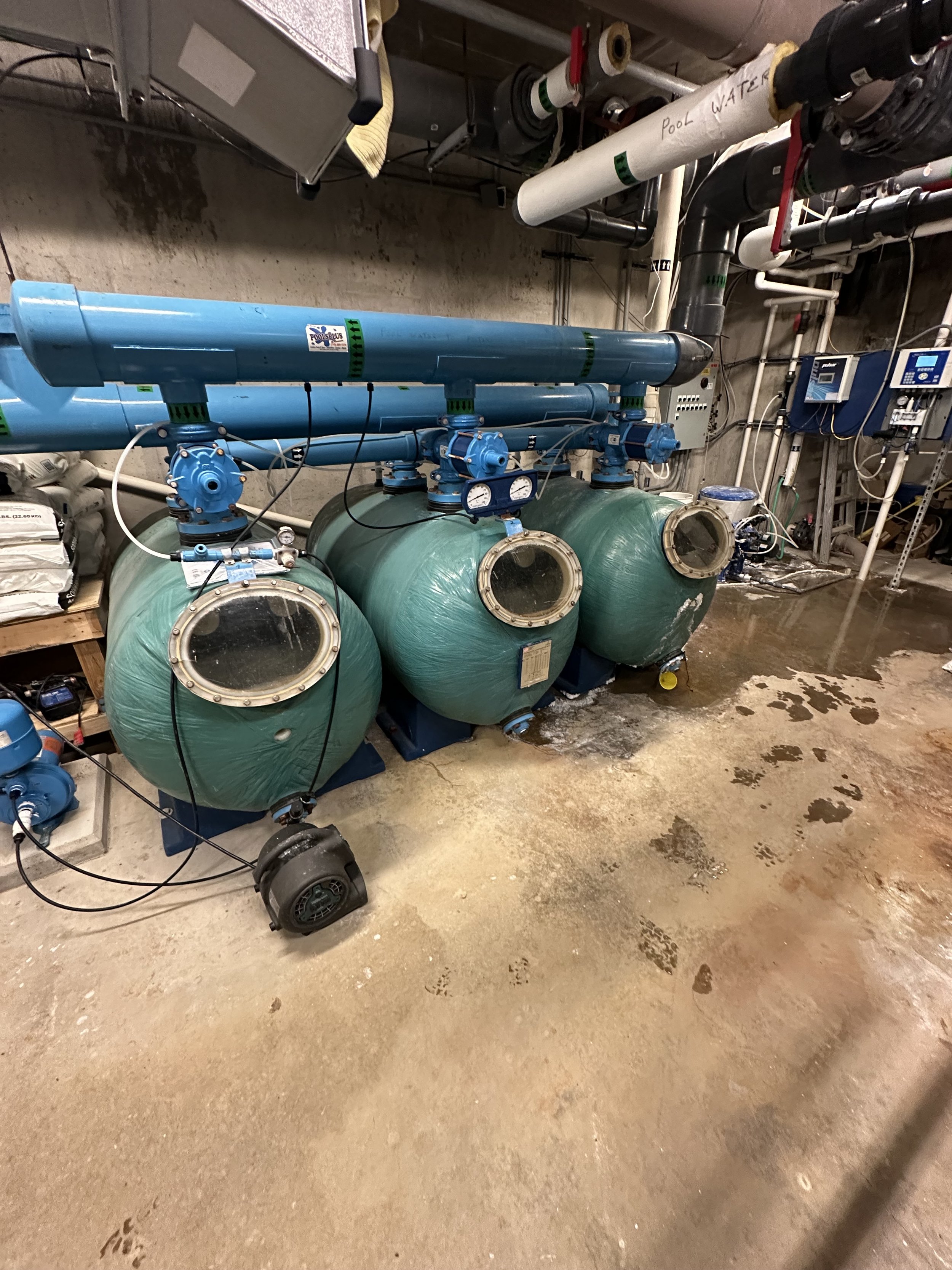 Commercial pool filtration system with green sand filter tanks, blue PVC piping, pumps, valves, and control panels in a basement mechanical room