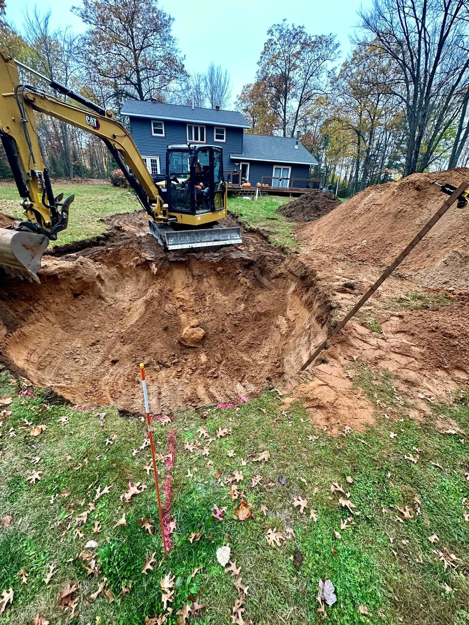 Compact CAT excavator digging a large trench in the backyard of a dark blue house, with piles of soil and autumn trees.
