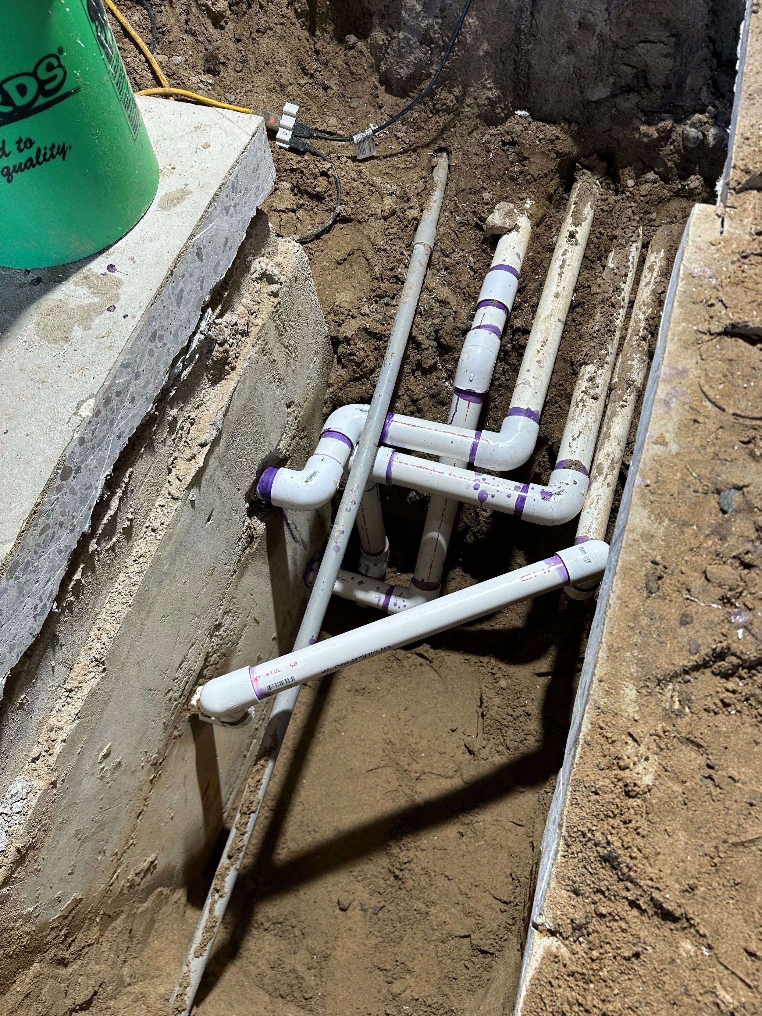 Exposed white PVC plumbing pipes with purple joints installed in a dirt trench beside a building foundation.