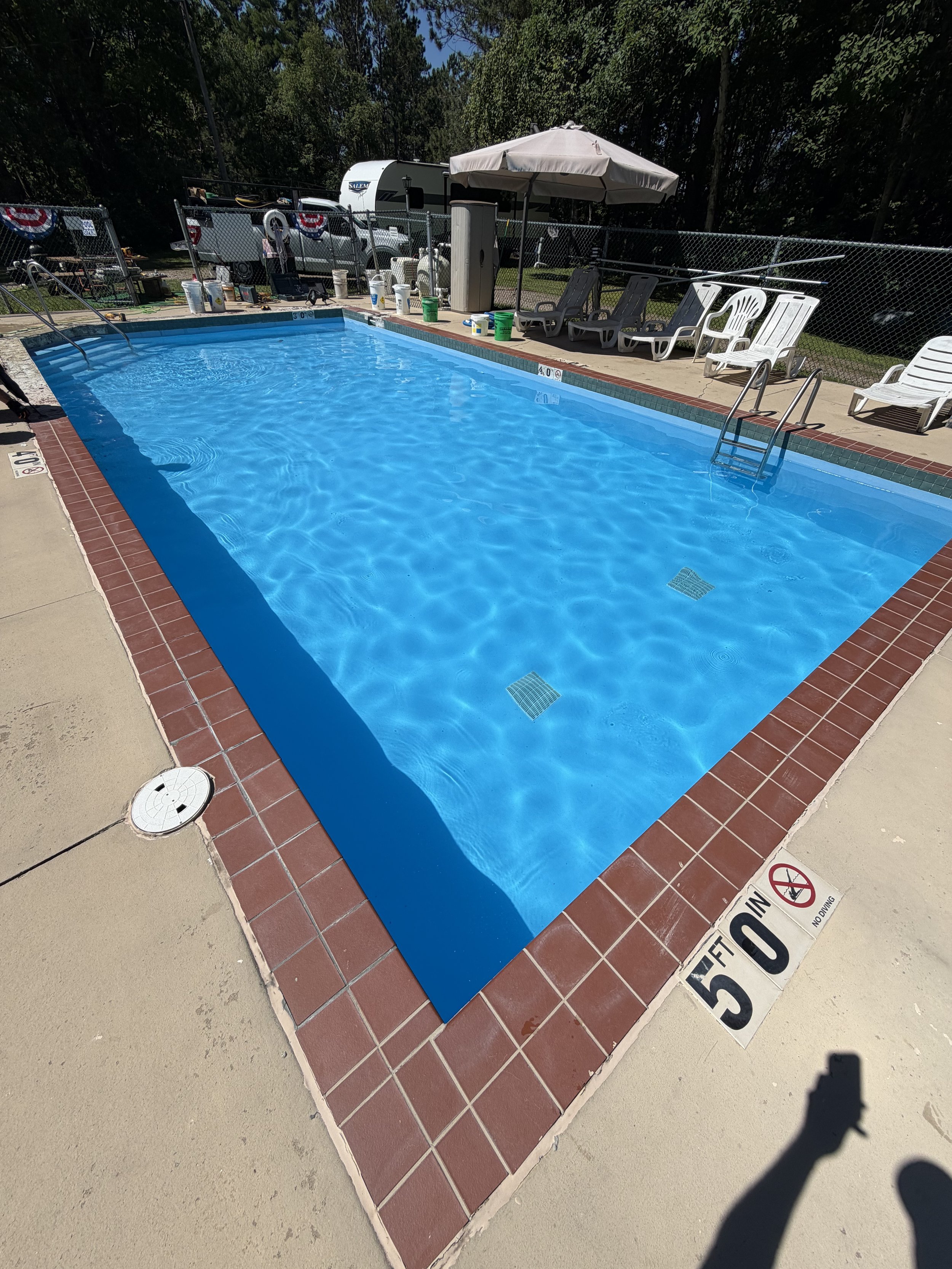 Outdoor swimming pool with lounge chairs, umbrella, maintenance supplies, and a Salem travel trailer behind a chain-link fence