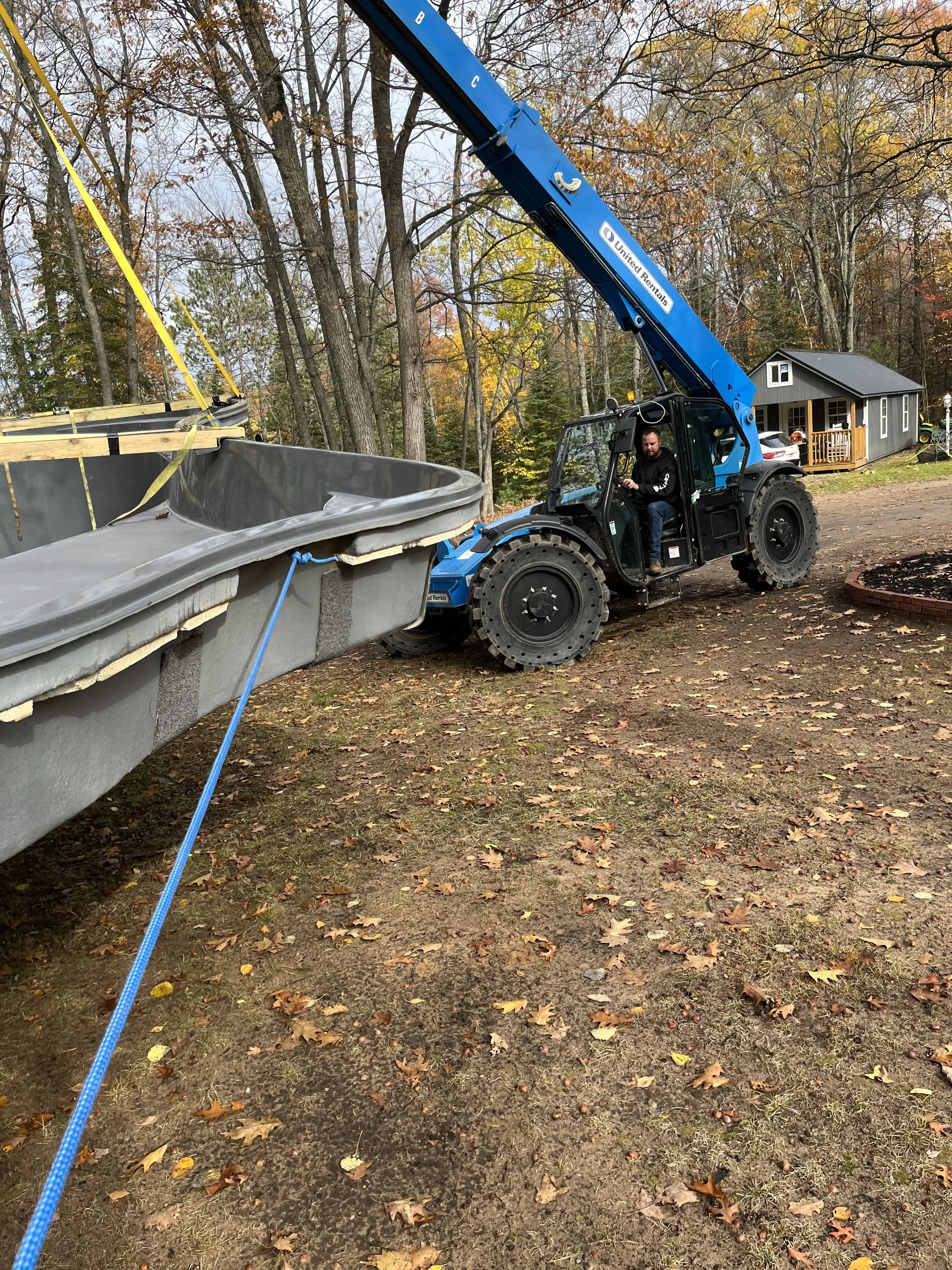 Blue telehandler lifting a large hot tub beside a wooded cabin in autumn