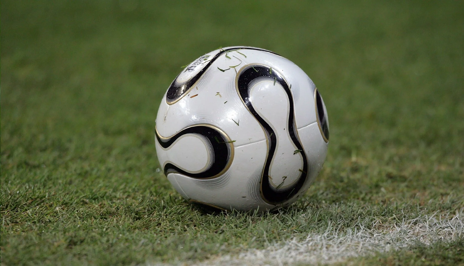 A white soccer ball, a 2006 adidas FIFA world cup ball,  with black and gold accents on a professional grass soccer field.