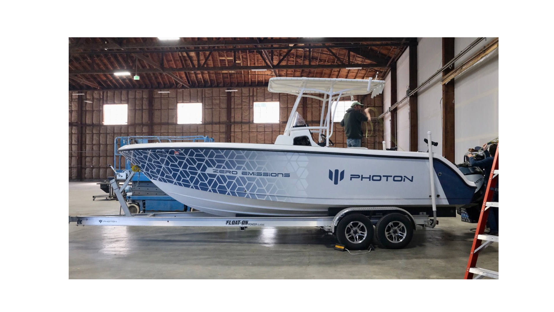 An electric boat named Photon with Zero Emissions on a trailer inside a warehouse, with a man working on it and another person seated at the back.