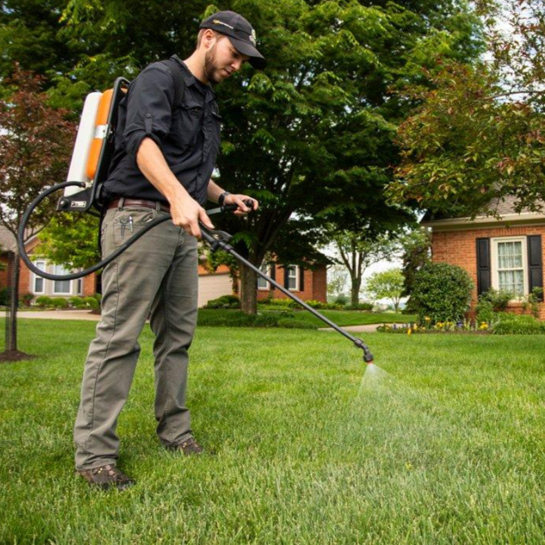 Man wearing black shirt, gray pants, and a black cap using a backpack sprayer to spray liquid on a lawn in a suburban neighborhood.