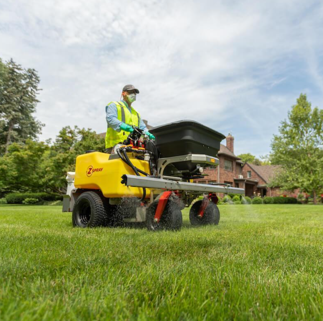 A person wearing safety gear and a vest operating a yellow spray machine in a grassy lawn, with a house and trees in the background.
