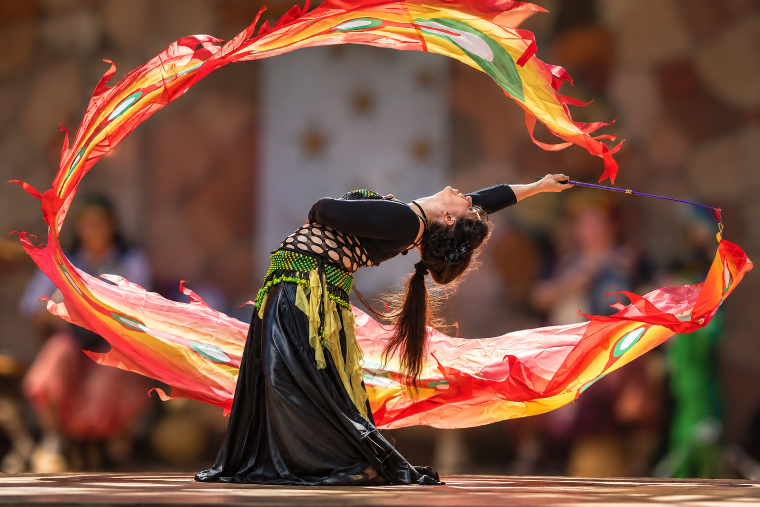 A woman performing a belly dance with vibrant orange, yellow, and green silk veils, kneeling on a stage during a cultural festival.