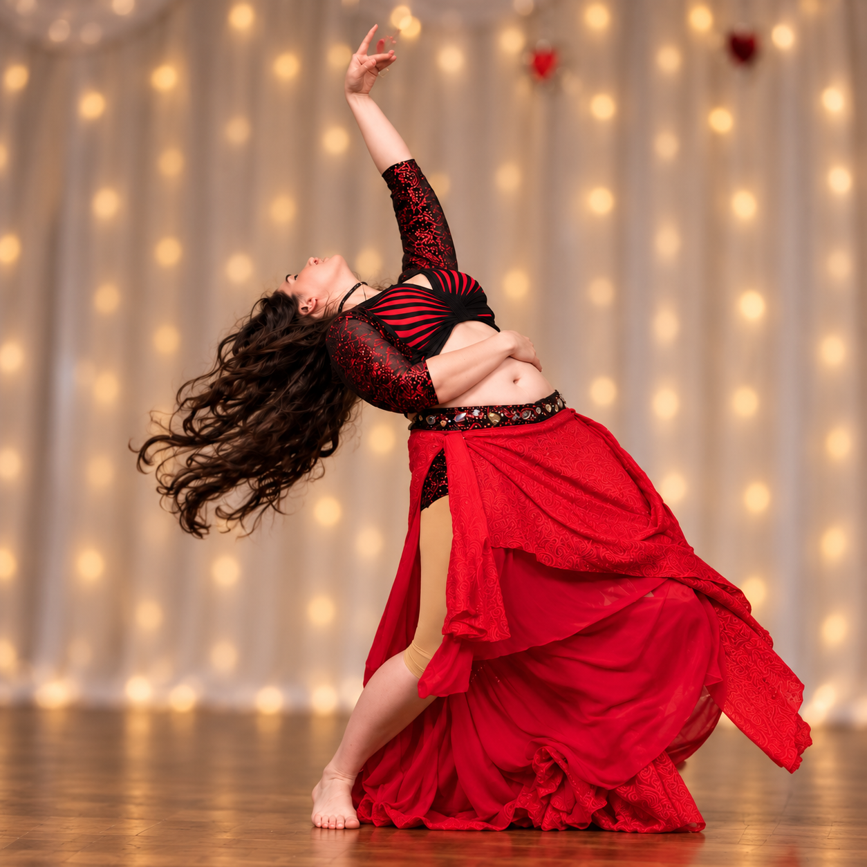 A woman in a red and black dance costume performs a graceful belly dance pose on stage, with a warm, decorated curtain background with lights.