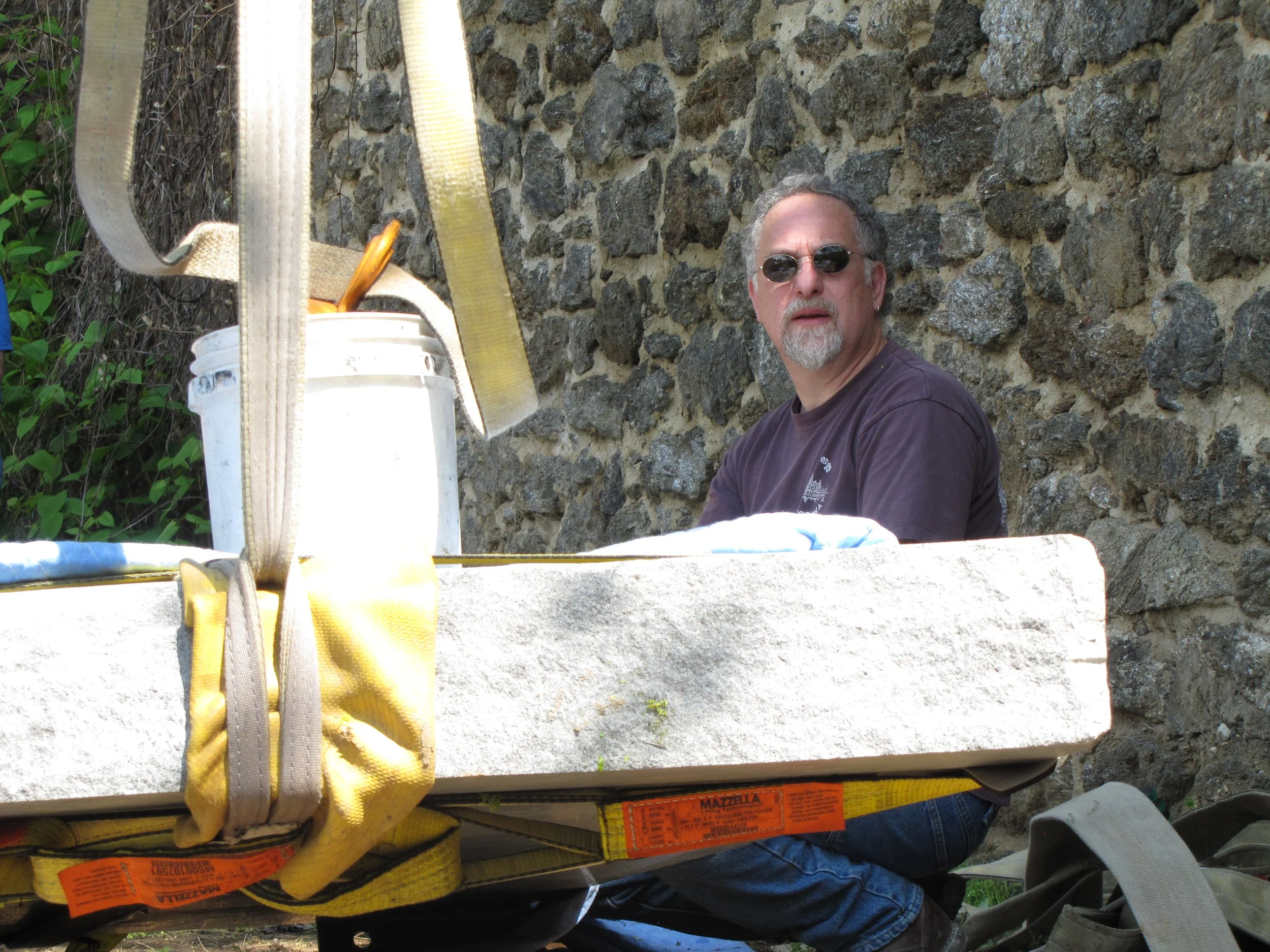 A man wearing sunglasses sitting in front of a stone wall, with a yellow safety harness and a white bucket in the foreground.