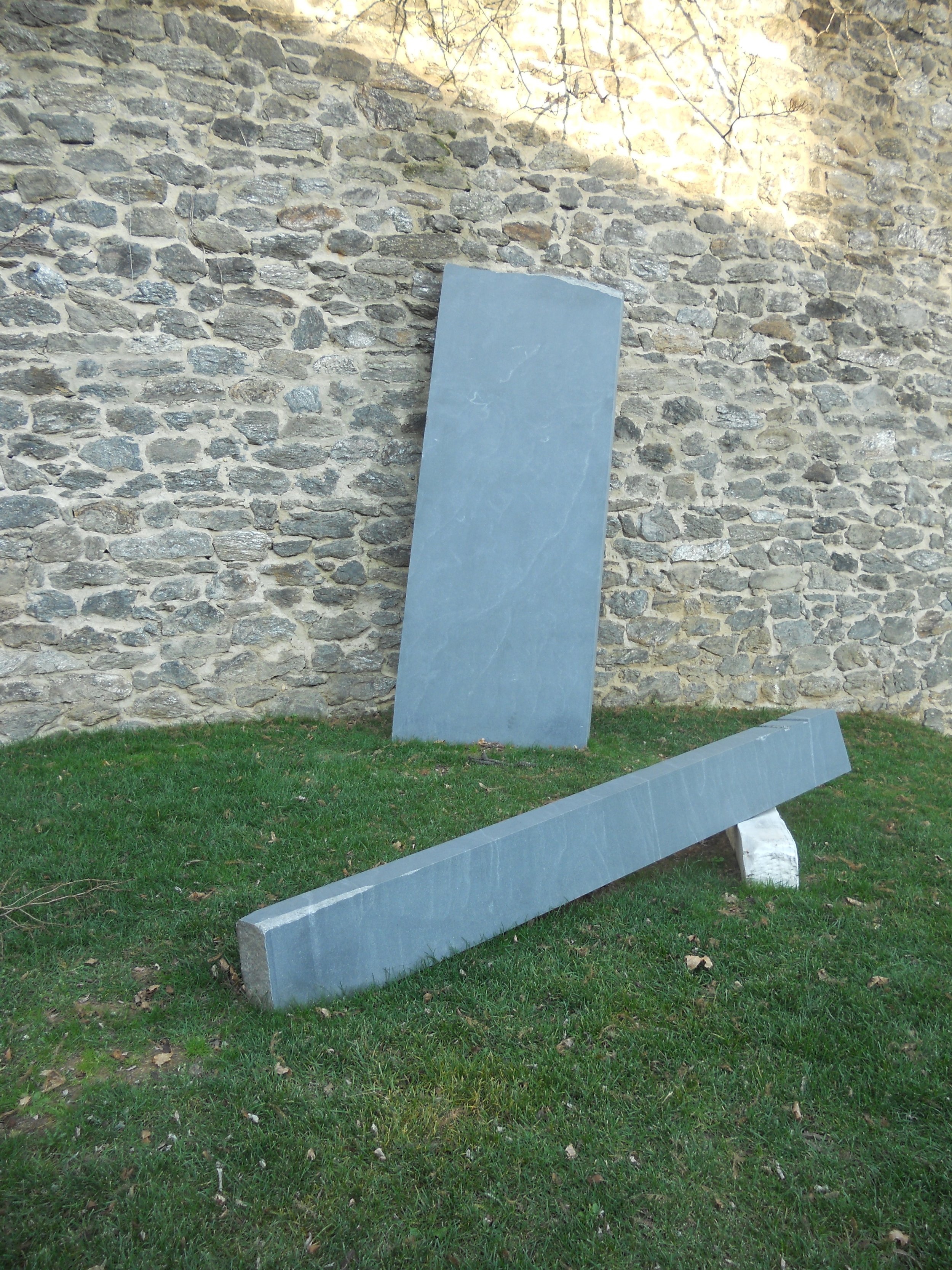 Three large, rectangular gray stone slabs leaning against a stone wall, with one slab on the ground and another slightly tilted, on grassy ground with fallen leaves.