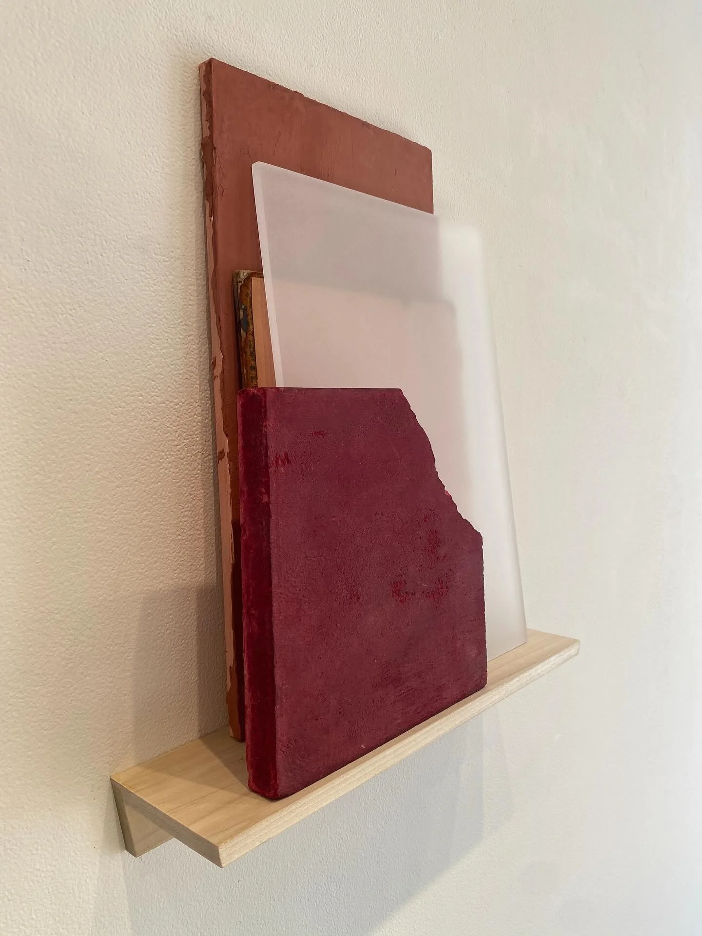 Three wooden shelves mounted on a white wall, with the middle shelf holding a white rectangular object, and the bottom shelf holding a red textured rectangular object.