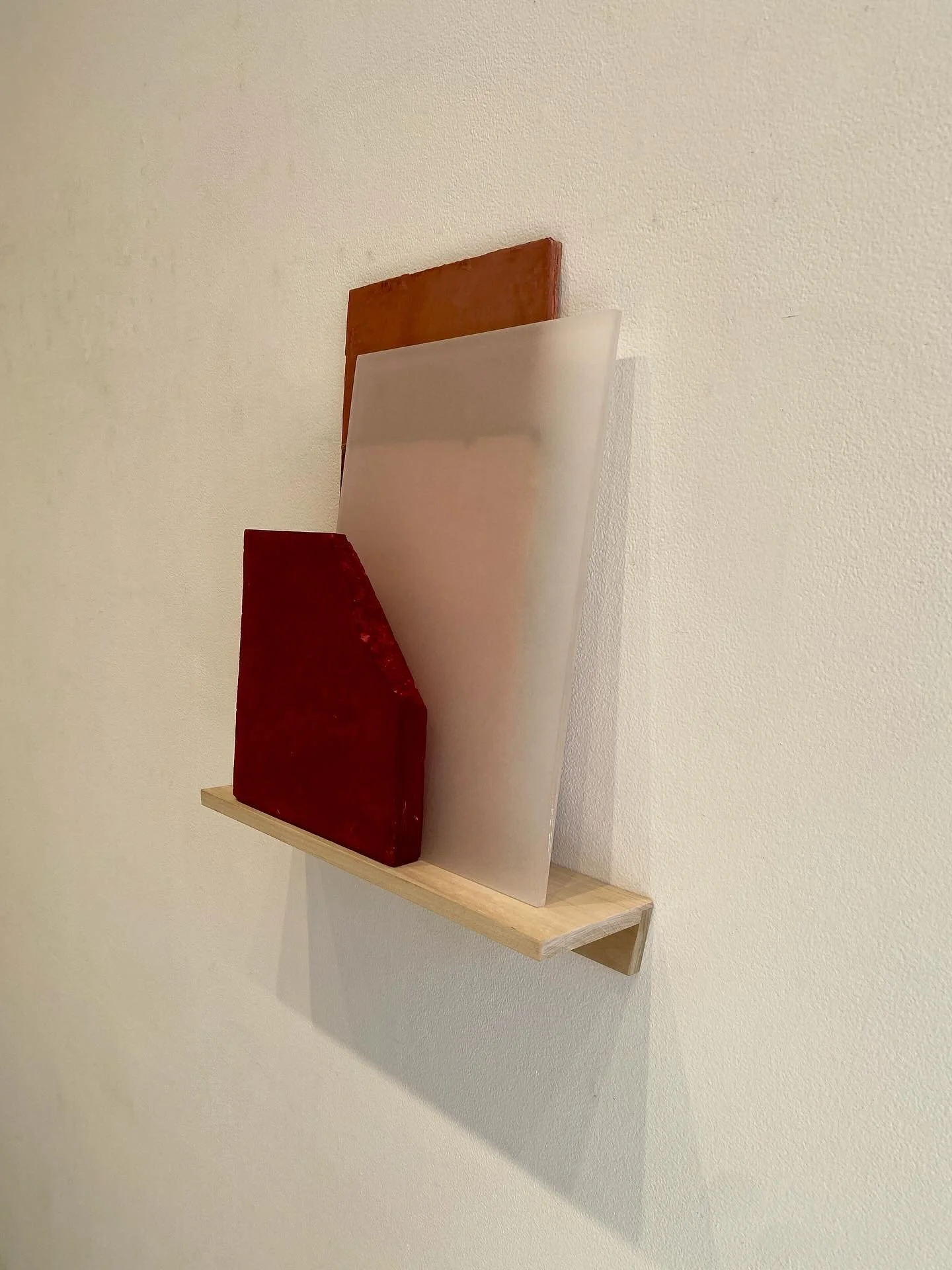 Abstract sculpture composed of three rectangular blocks in red, white, and brown mounted on a wooden shelf against a beige wall.