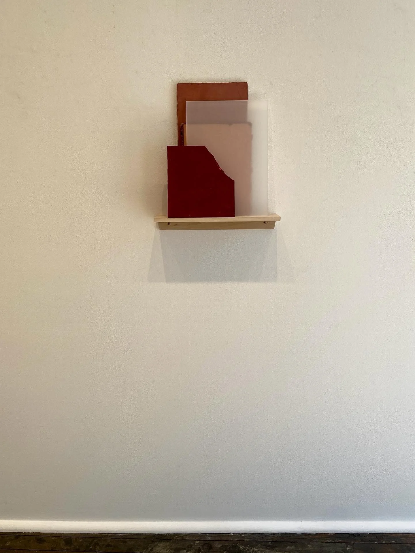 A small wooden shelf mounted on a plain wall with several wooden and frosted glass pieces, some painted in red, arranged vertically on the shelf.