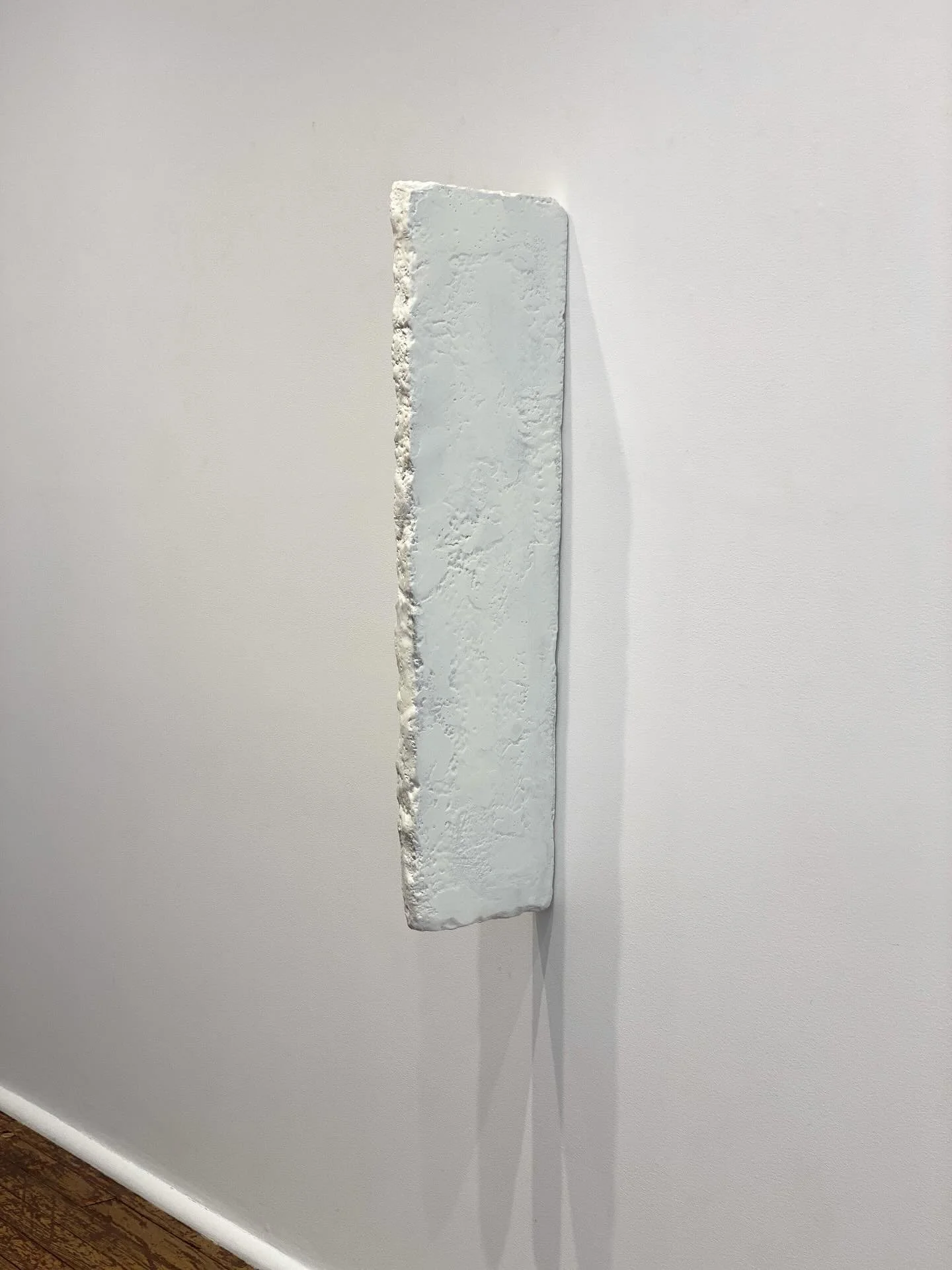 A thin rectangular sculpture mounted on a white wall, with a textured uneven surface and irregular edges.